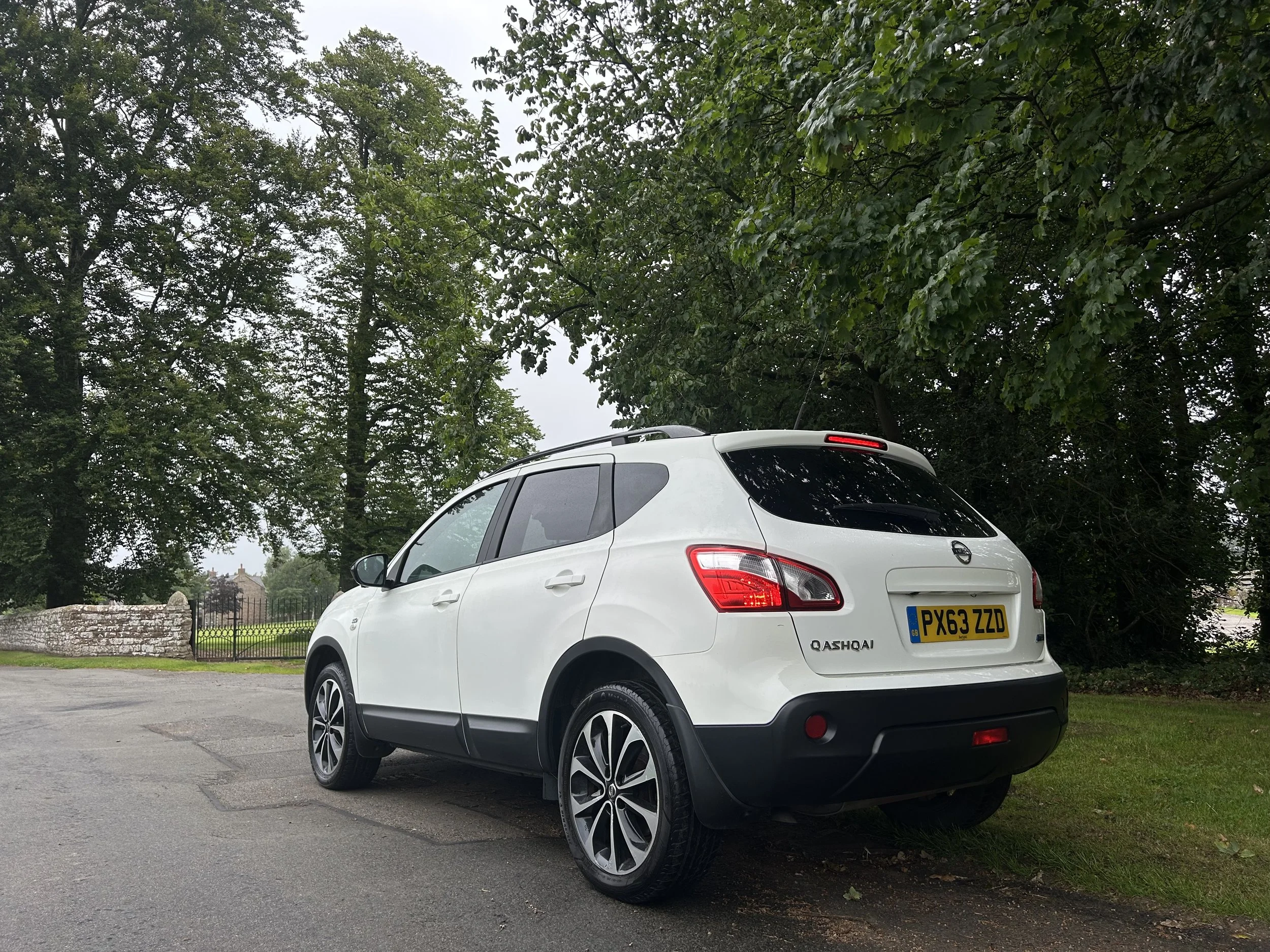 White Nissan Qashqai parked near green trees and a stone fence in a park setting.