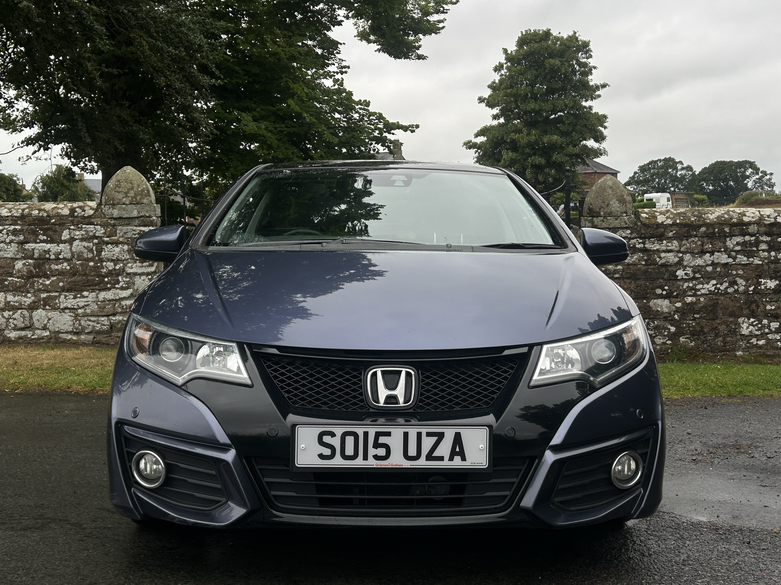 Front view of a dark gray Honda car with a UK license plate parked in front of a stone wall and trees, on a cloudy day.