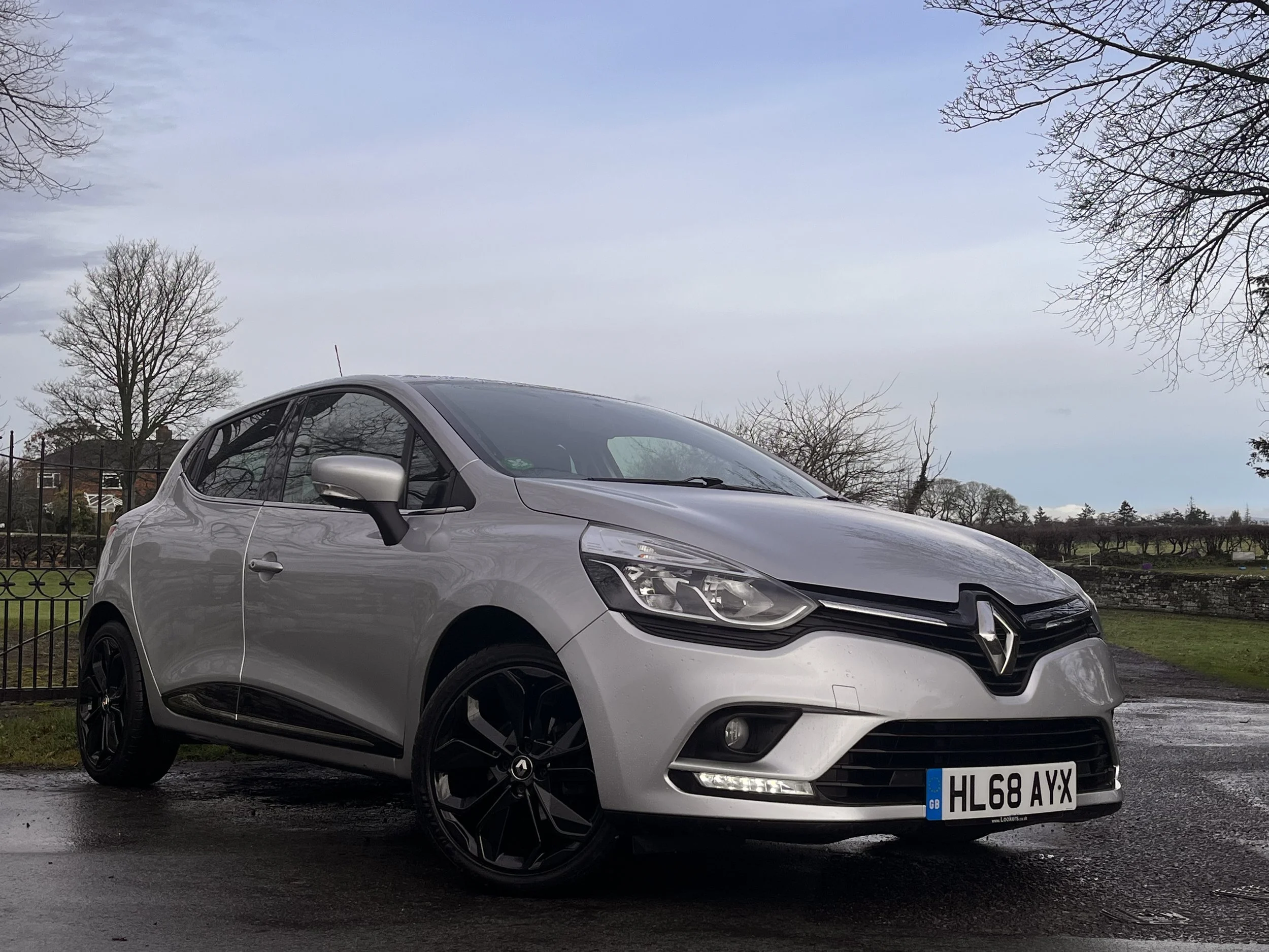 A silver Renault hatchback car parked outdoors on a paved wet surface with a fence and leafless trees in the background, under cloudy sky.