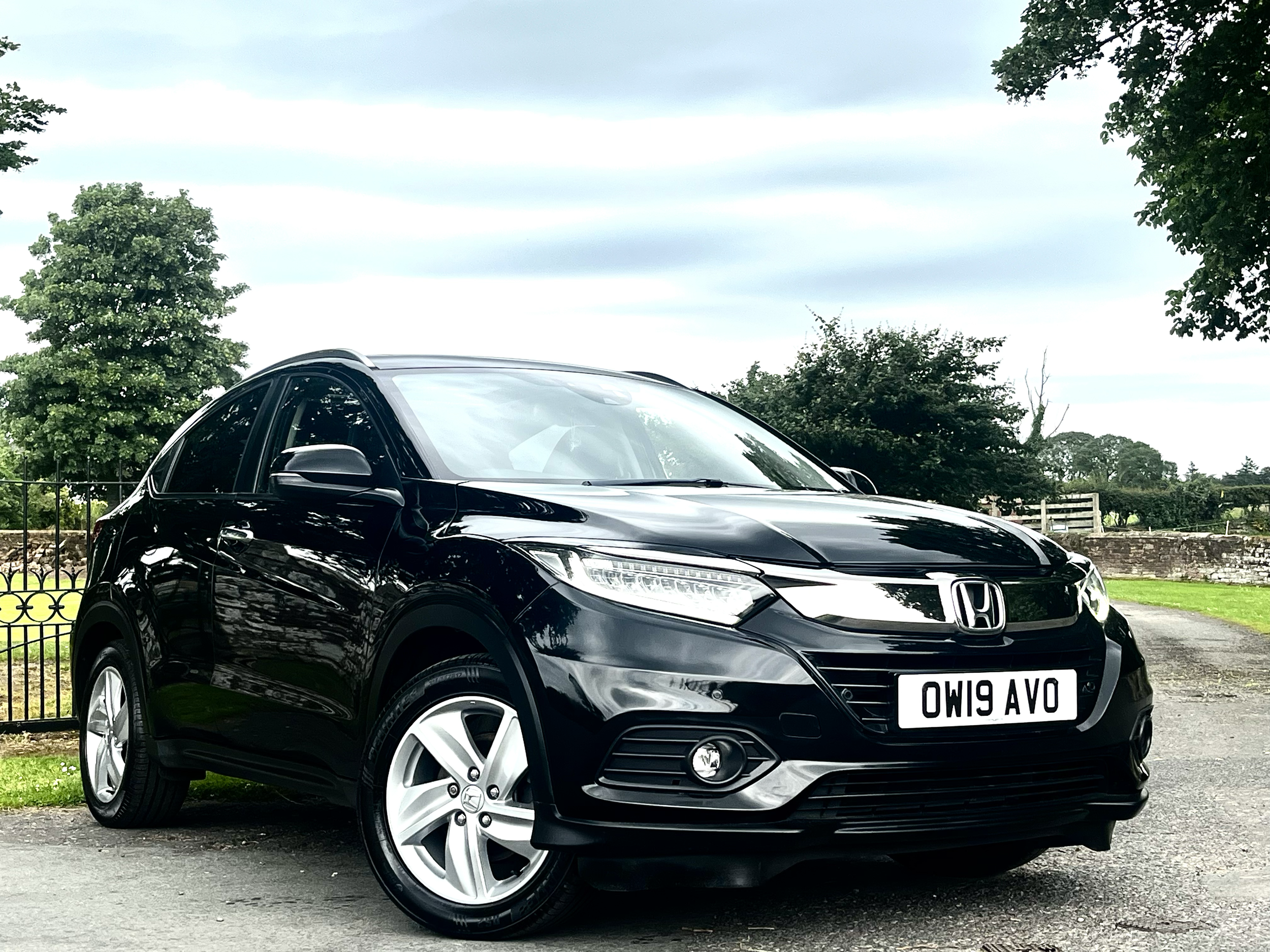 A black Honda car parked outdoors with trees and a cloudy sky in the background, displaying a white license plate with the text "OZVO 619W".