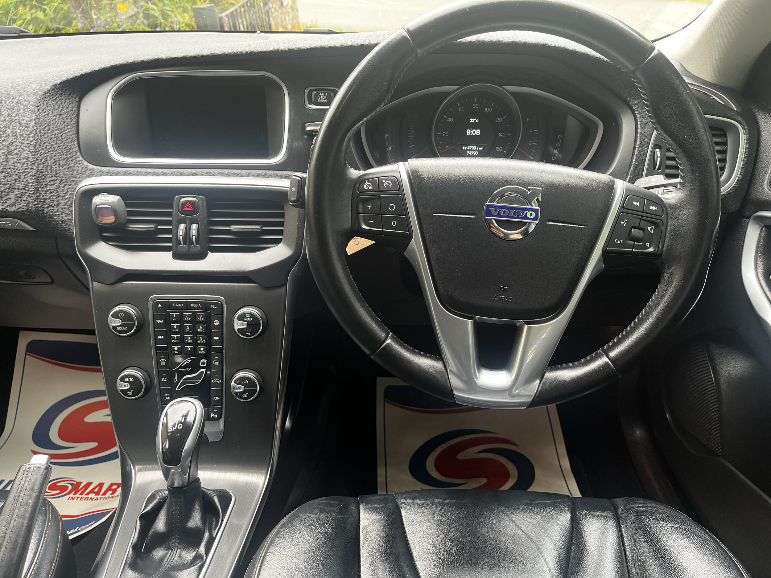 Interior of a showroom car dashboard and steering wheel with a Volvo emblem, digital speedometer, and controls, with a partial view of the outside through the windshield.