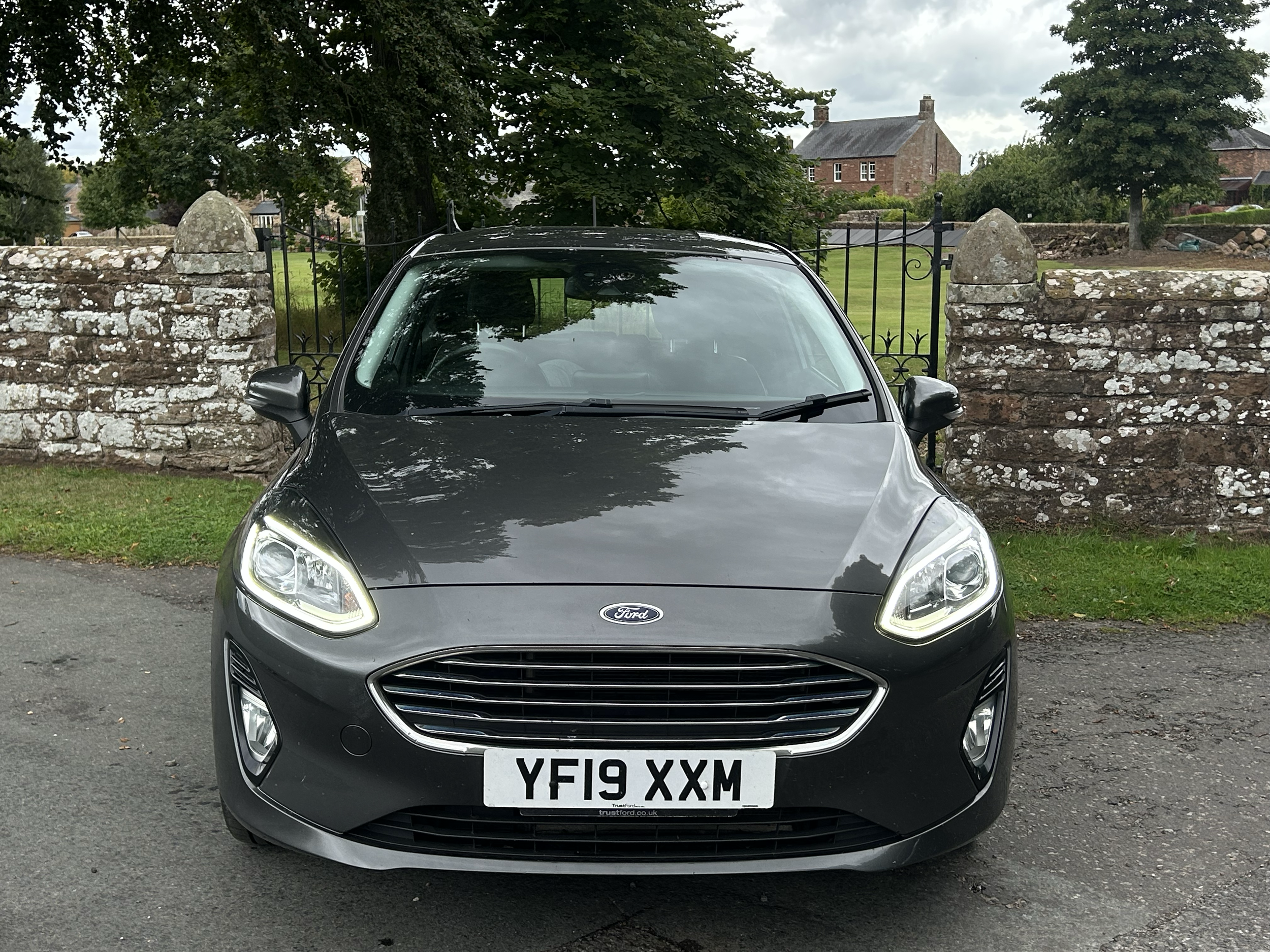 Black Ford vehicle parked in front of an old stone wall and a wooden gate, with green trees and historic brick buildings in the background.