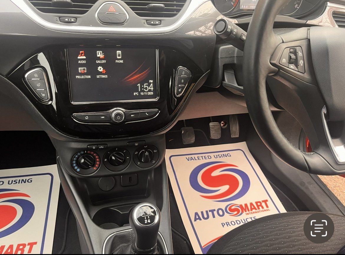 Interior view of a car dashboard showing a touchscreen display, climate control knobs, manual gear shift, and steering wheel, with floor mats labeled AUTO SMART INTERNATIONAL.