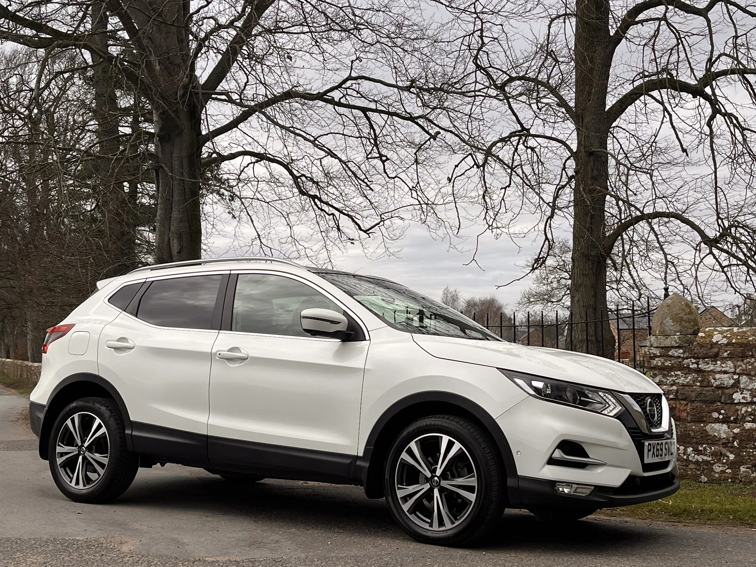 White Nissan SUV parked on a street with leafless trees and a stone wall in the background.