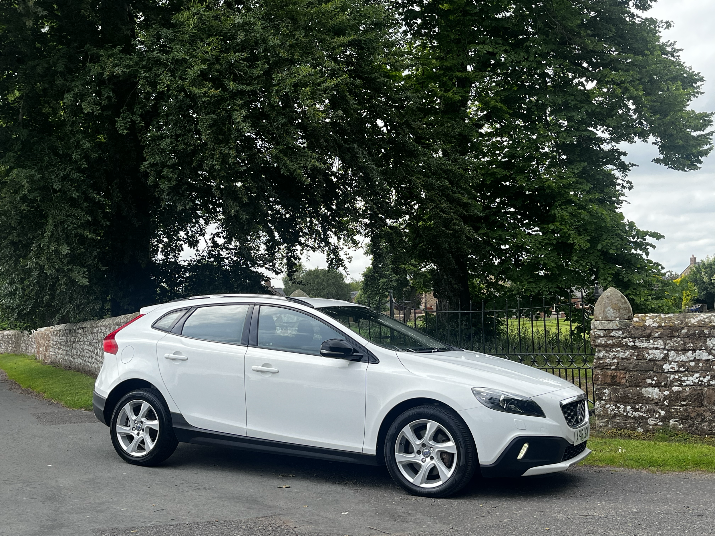 A white modern compact hatchback car parked on the side of a street in front of a short stone wall and a green metal fence. Behind the fence, there are large trees with green foliage and a partly cloudy sky.