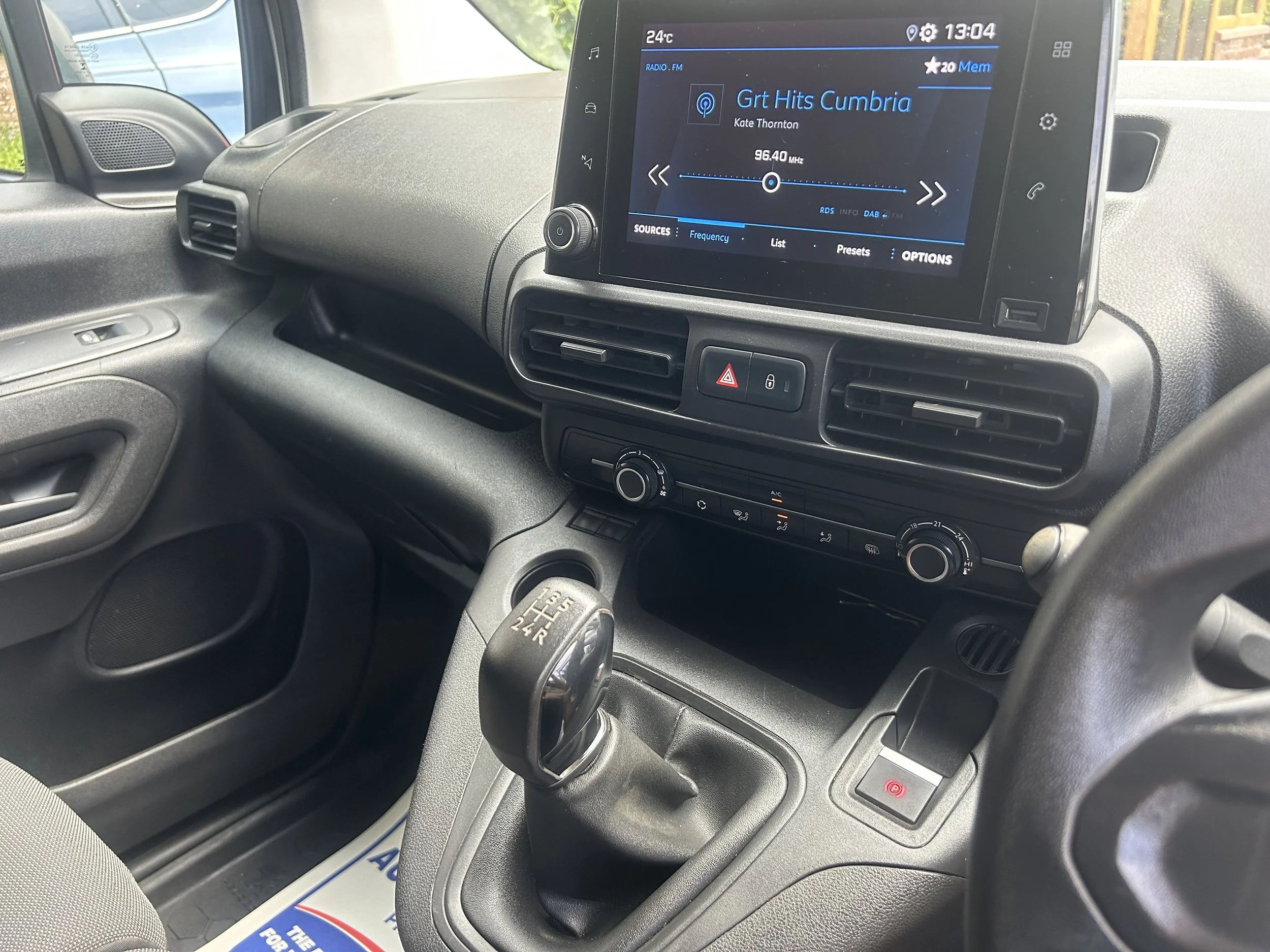 Inside the front cabin of a vehicle, showing a dashboard, a gear shift, and a digital infotainment screen playing a radio station