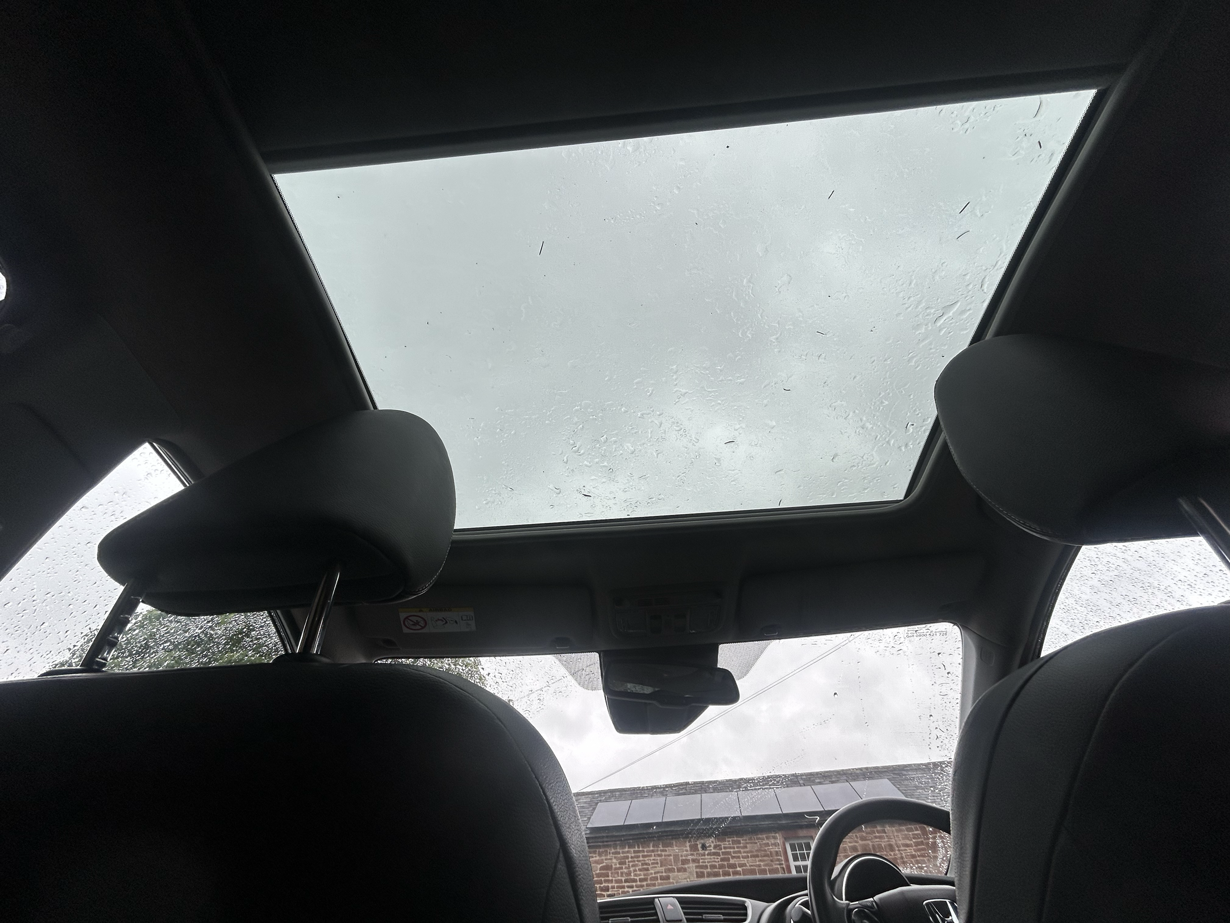 View of a car's interior showing a sunroof with raindrops on the glass, seats, and part of a dashboard, with a building and electrical wires visible outside through the sunroof.