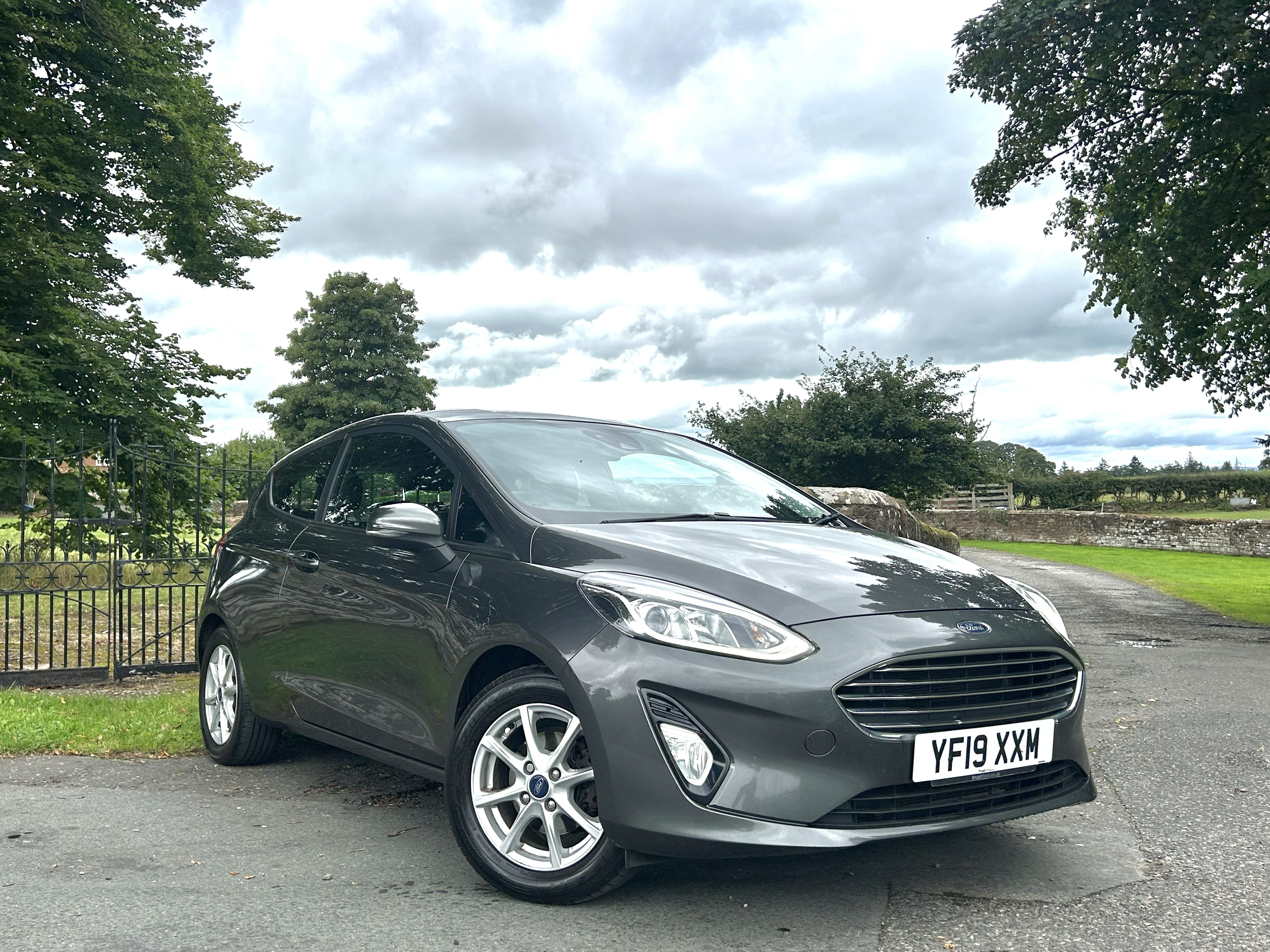 A gray Ford car with a British license plate parked on a paved surface, surrounded by trees and a cloudy sky.