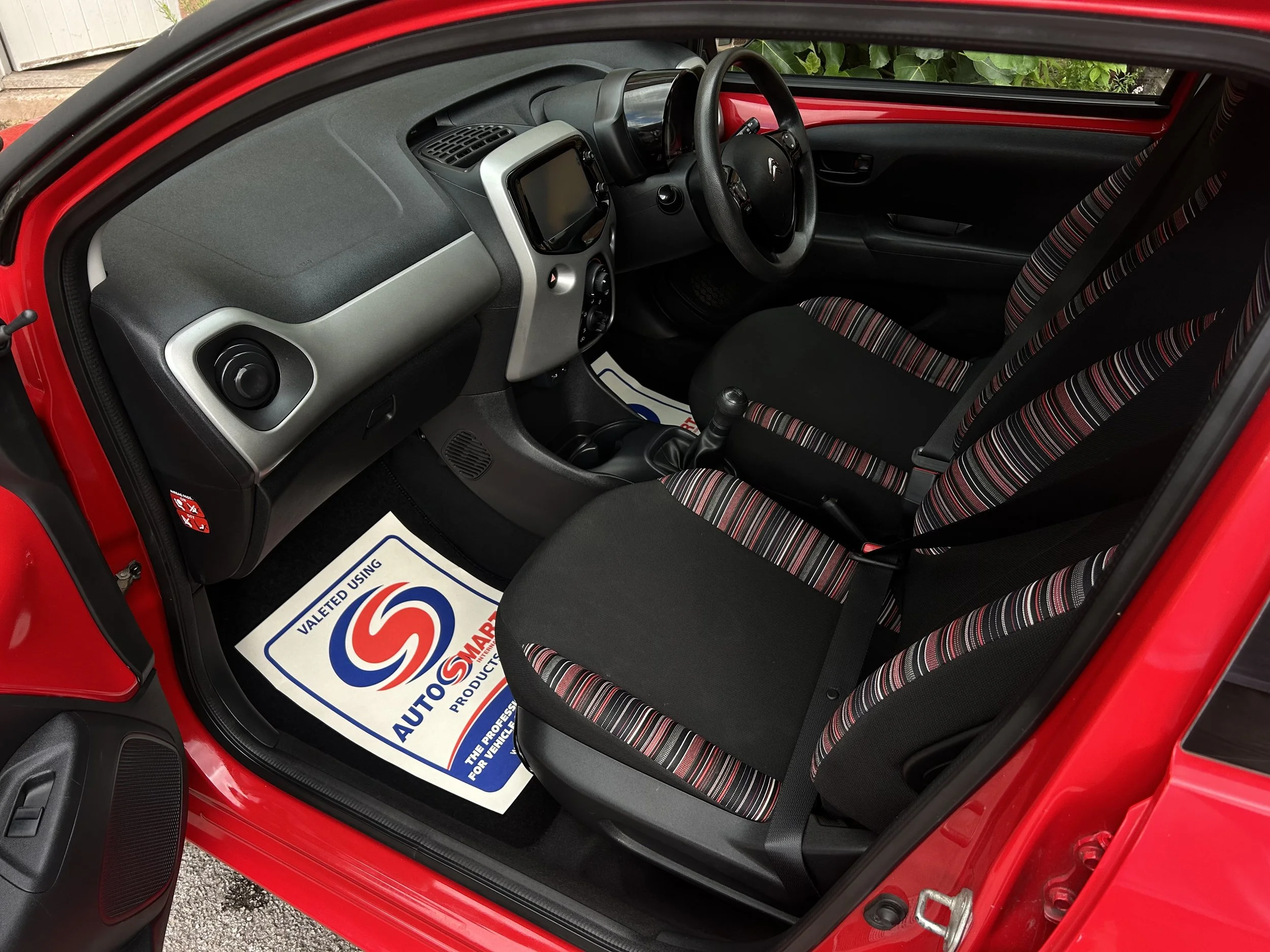 Interior of a red compact car showing the dashboard, steering wheel, gear shift, and front seats with striped fabric upholstery.