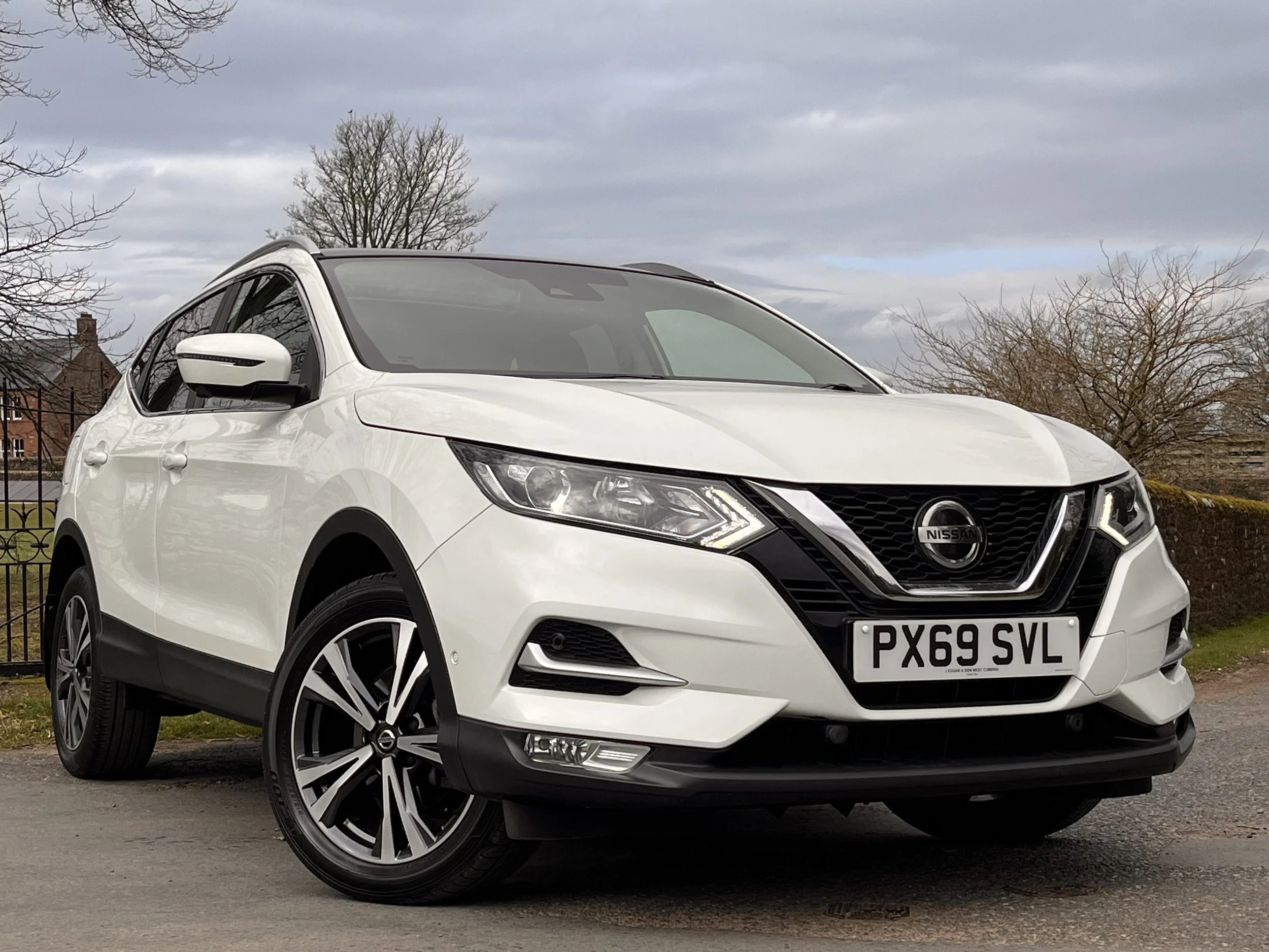 White Nissan SUV parked on a street with a cloudy sky and leafless trees in the background.