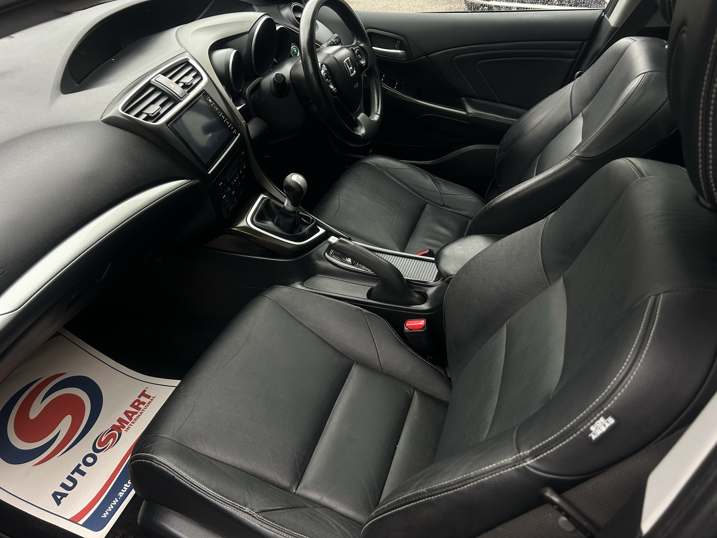 Interior of a black car showing the driver's seat, steering wheel, dashboard, and gear shift, with a paper mat on the floor that says 'Auto Mart'.