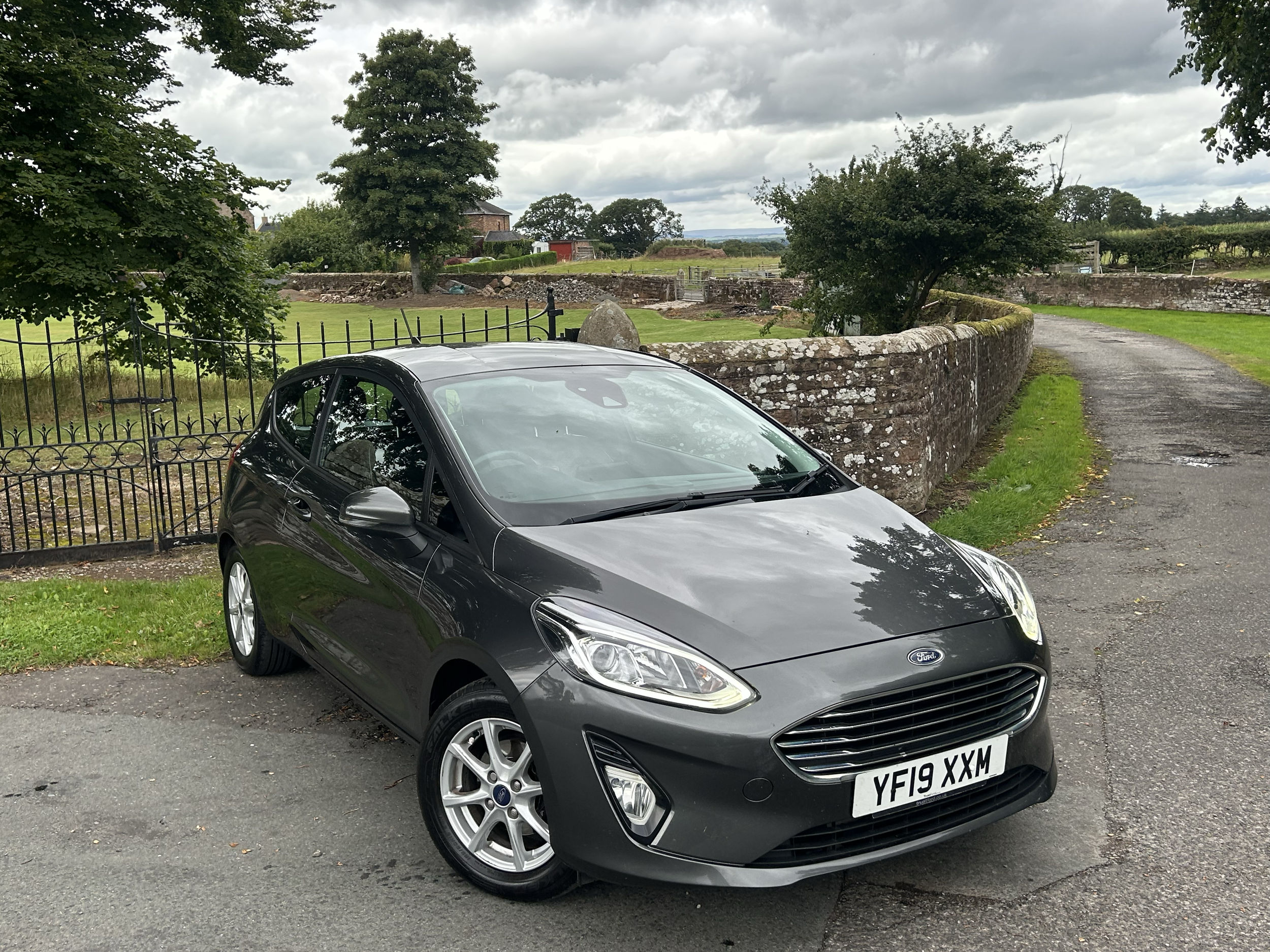 A black Ford car parked on a paved road next to a stone wall and a grassy area. There are trees and a cloudy sky in the background.