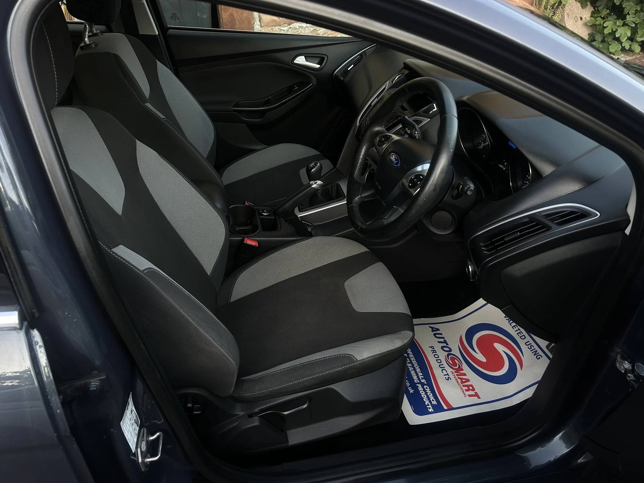 Interior of a black Ford car showing front seats, steering wheel, dashboard, gear shift, and floor mat with a logo.