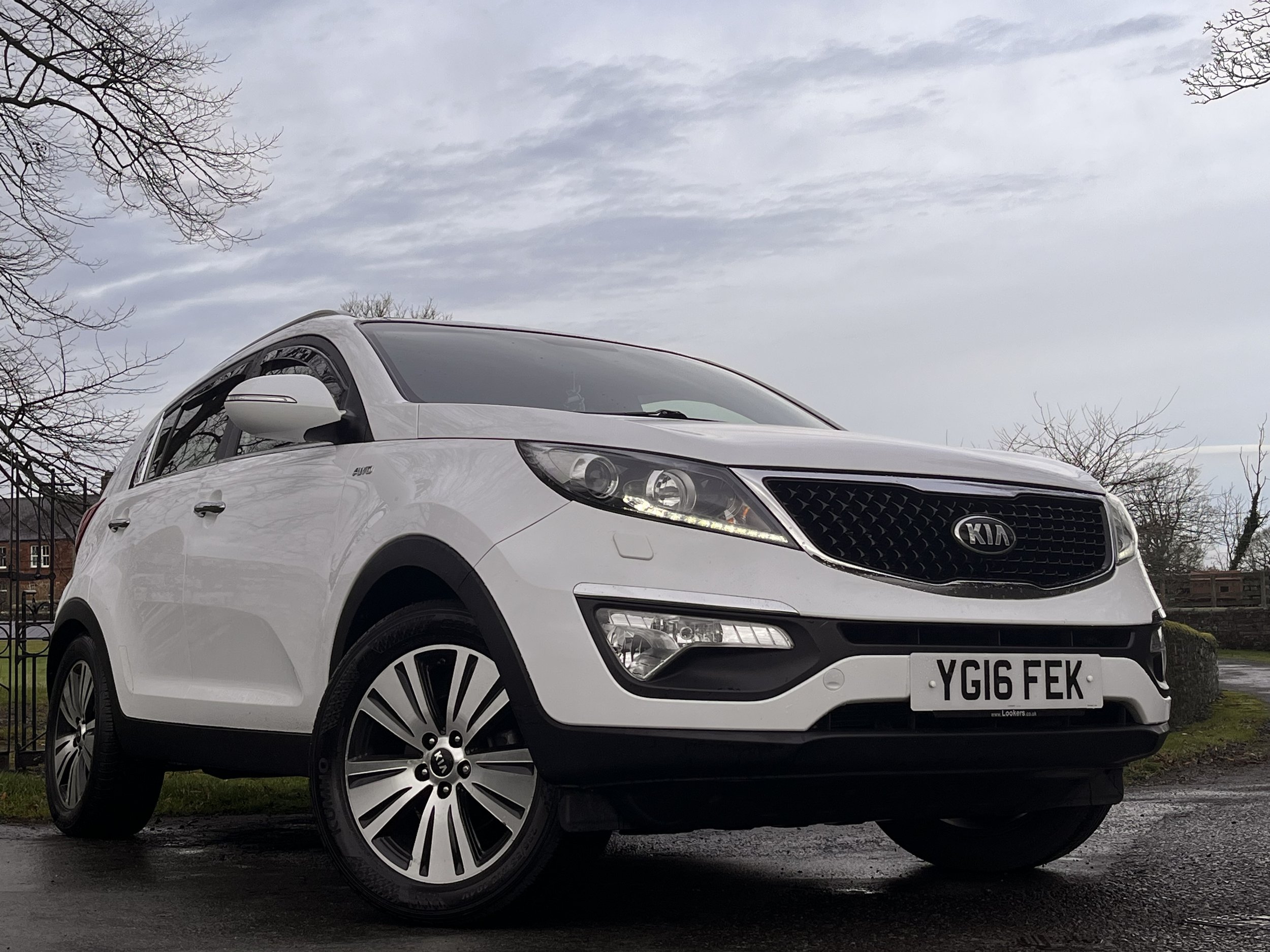 White Kia SUV parked on a wet surface with trees and a cloudy sky in the background.