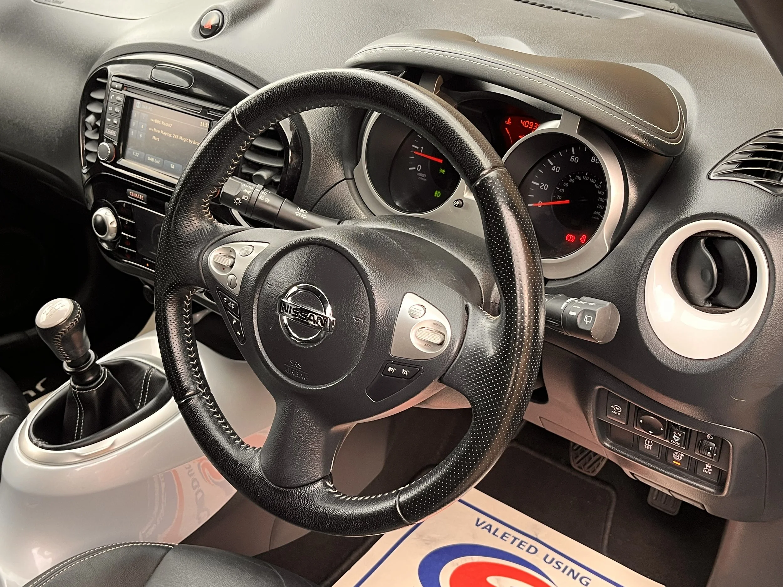 Interior of a Nissan vehicle showing the steering wheel, dashboard with speedometer and other gauges, infotainment system, gear shift, and various control buttons.