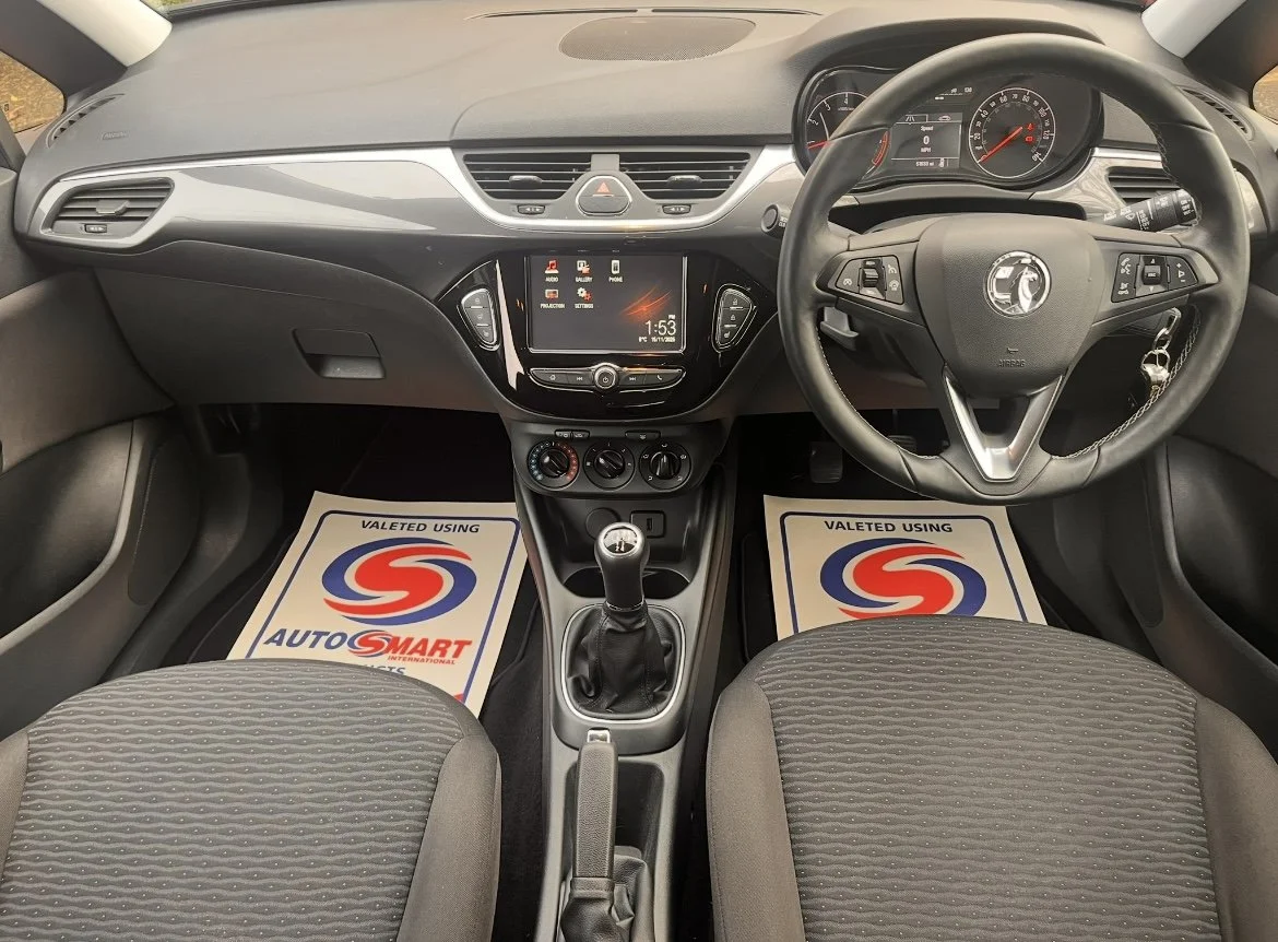 Interior of a car showing the dashboard, steering wheel, gear shift, and front seats. The car has a central display, air vents, and control knobs. Floor mats with the AutoSmart logo are visible.