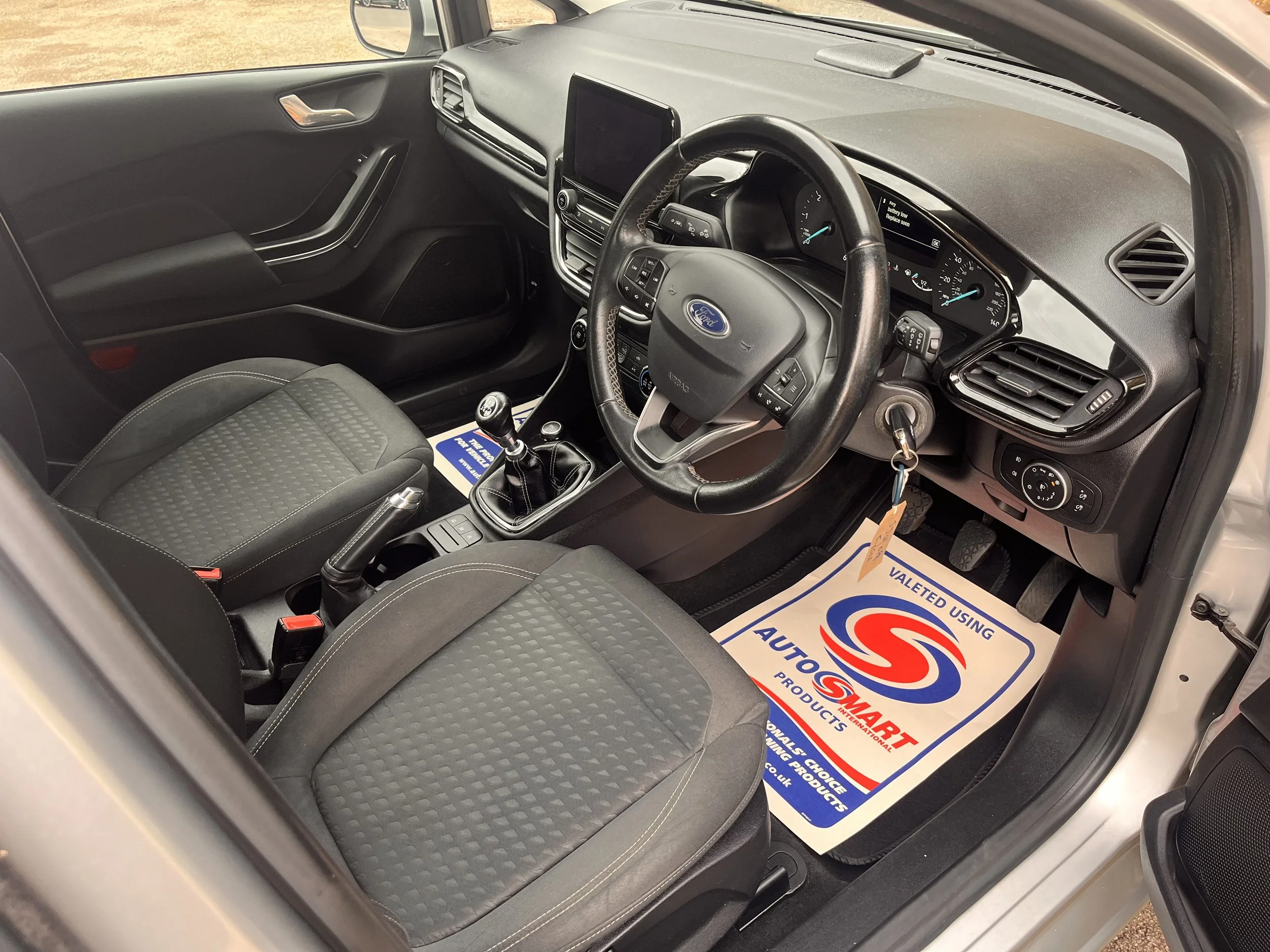 Interior view of a Ford vehicle showing the front seats, dashboard, steering wheel, gear shift, and pedals, with an AutoSmart floor mat.