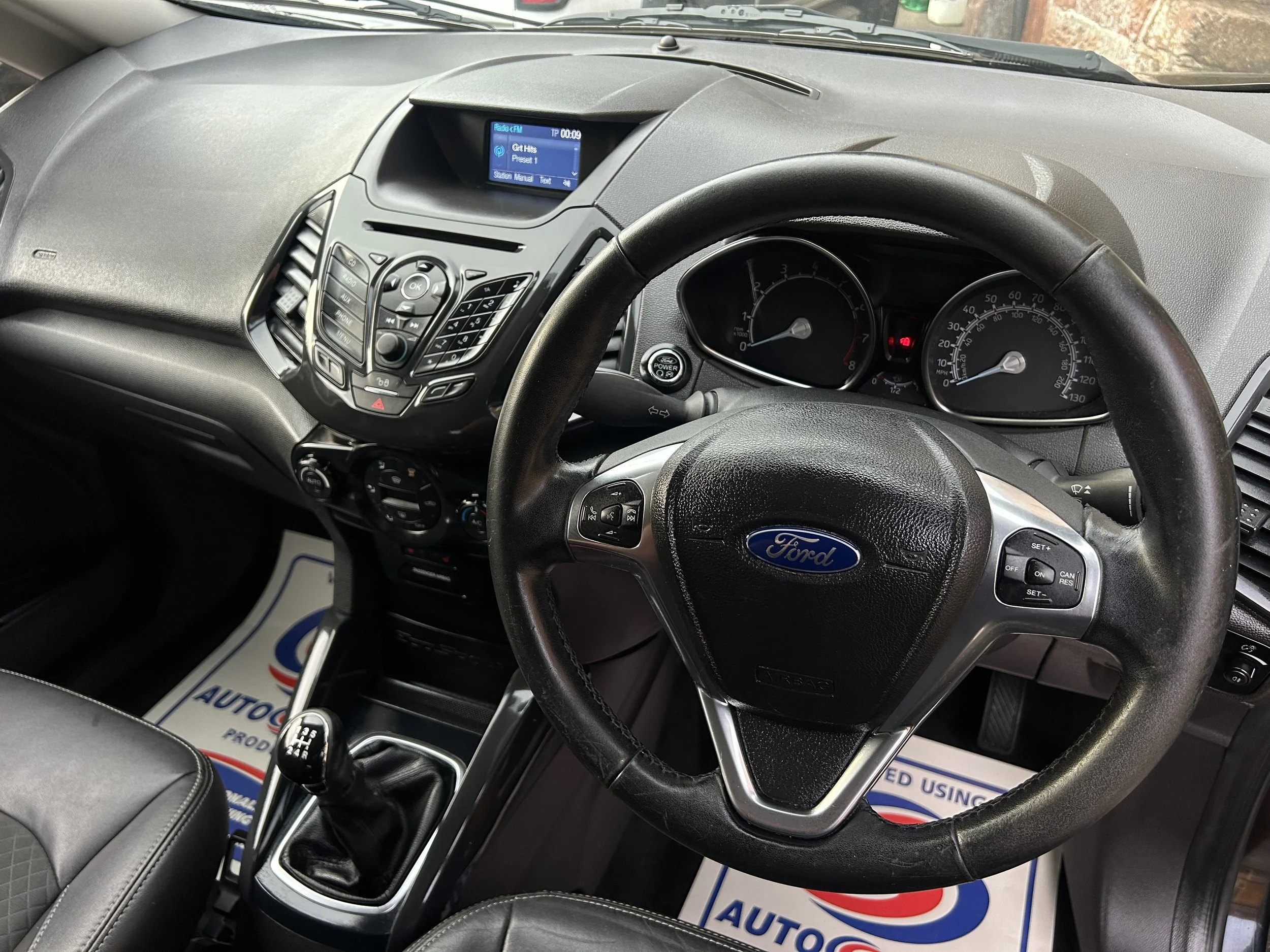 Interior view of a Ford vehicle dashboard, steering wheel, and gear shift lever with a paper floor mat underneath.
