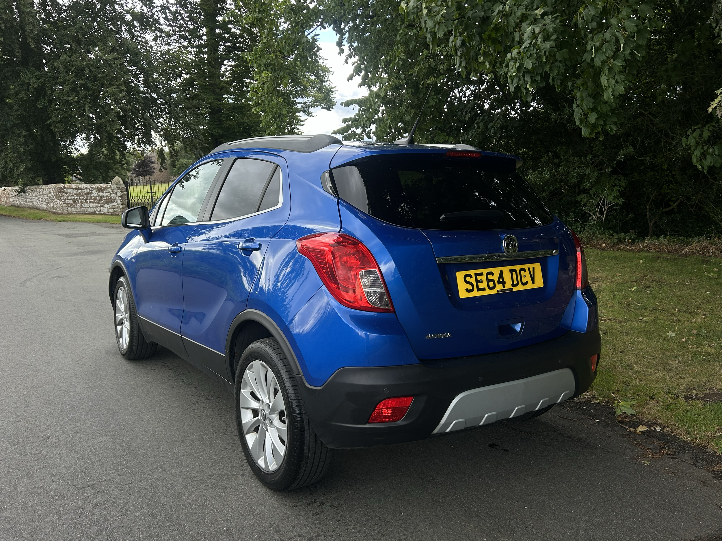Blue compact SUV parked on a street with a stone wall and green trees in the background.