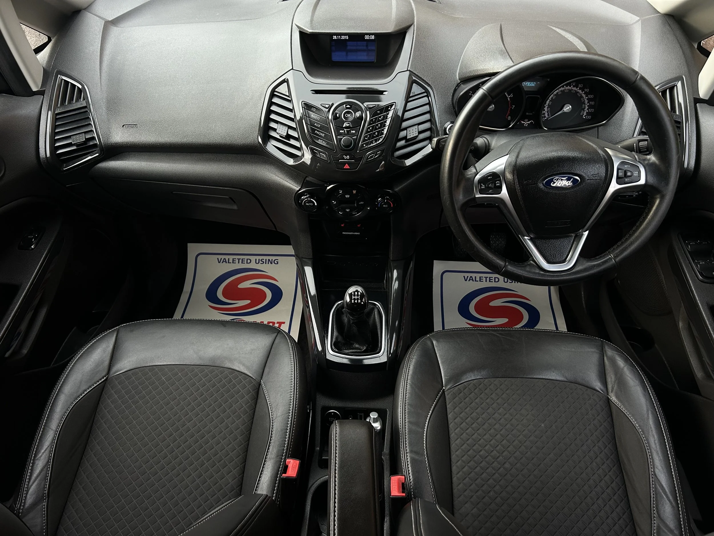 Interior of a Ford vehicle showing the dashboard, steering wheel, gear shift, and two black seats with diamond pattern inserts, with floor mats stating 'Valeted Using' and a logo.