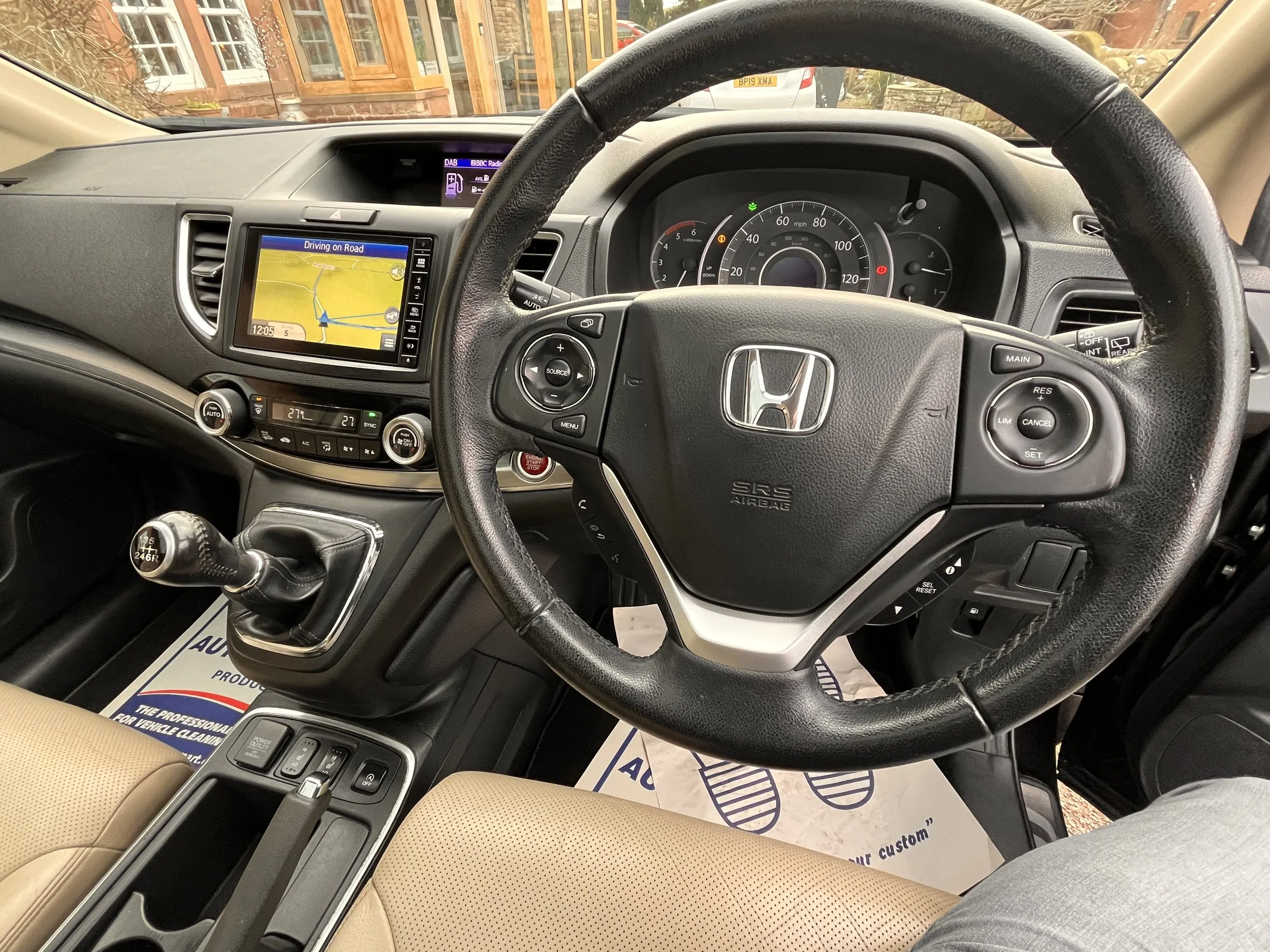 Interior of a Honda vehicle showing the steering wheel, dashboard with speedometer, multimedia screen, climate controls, gear shift, and beige seats.