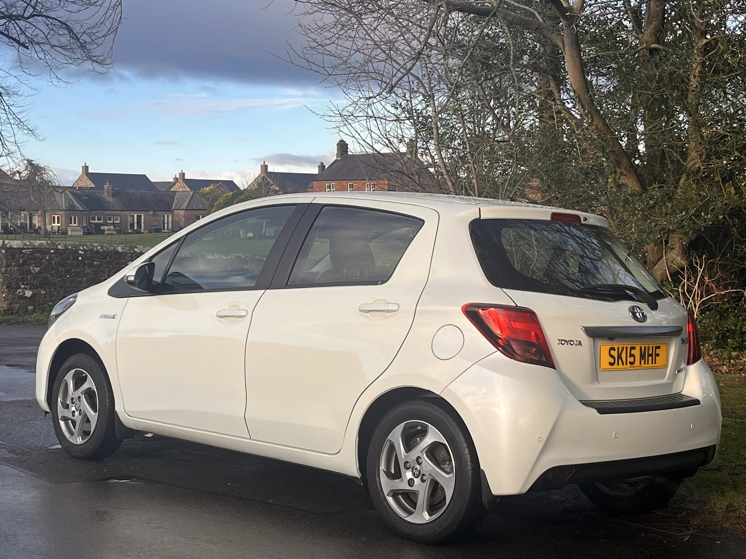 A white Toyota hatchback car parked on the side of a street with a brick wall and houses in the background. The car has a UK license plate reading 'SK15 MHF'.