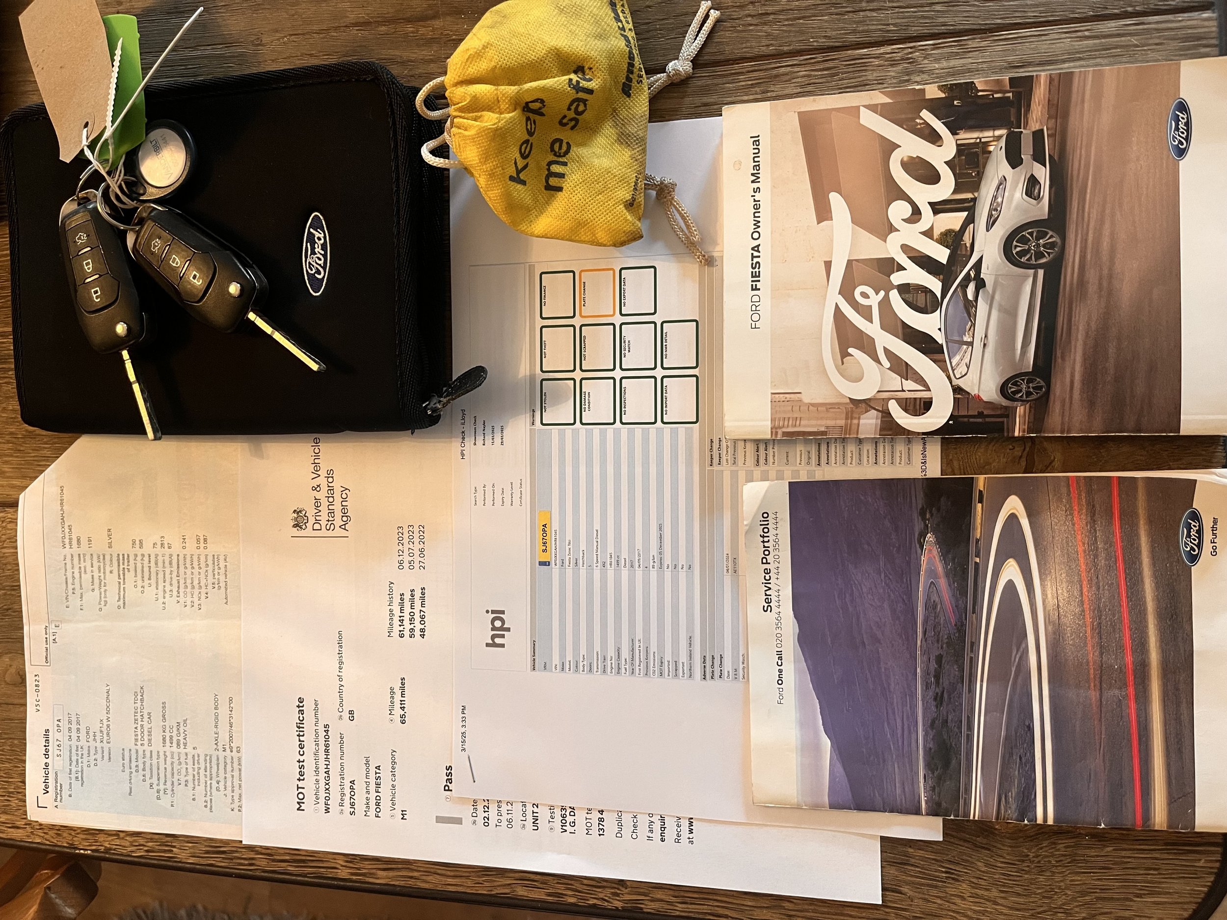 Car keys on a black Ford case, a yellow key pouch, a Ford Fiesta owner’s manual, and a Ford service portfolio booklet on a wooden table.