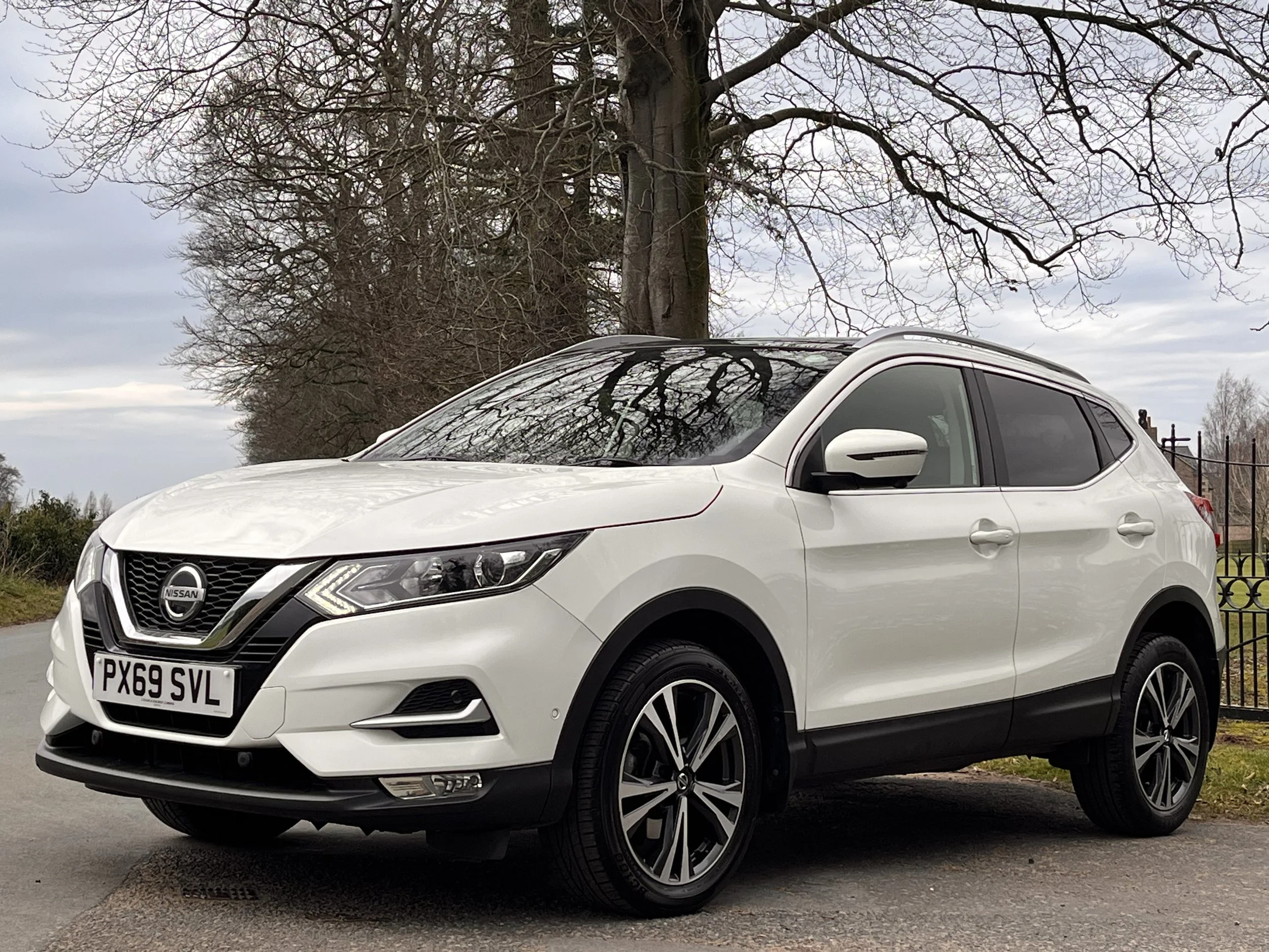White Nissan SUV parked on the side of a road with trees and a fence in the background, under an overcast sky.