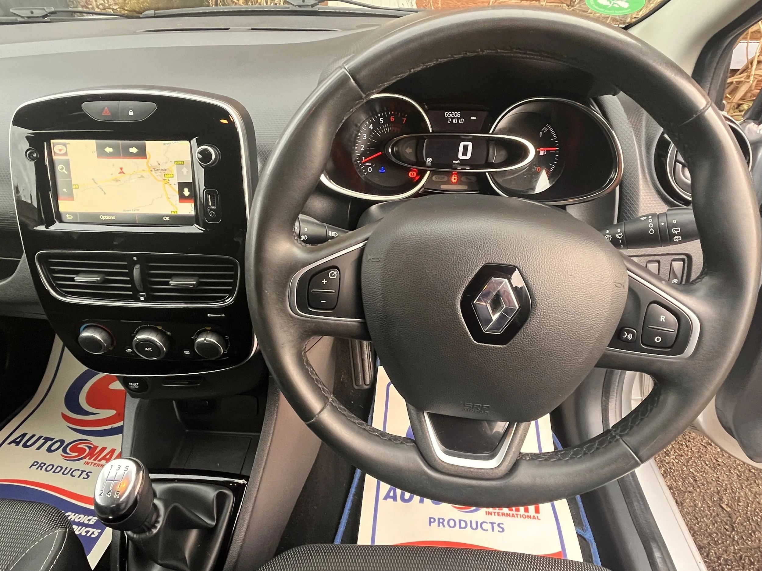Interior view of a Renault car dashboard featuring a steering wheel with Renault emblem, digital speedometer, navigation system screen, climate controls, and gear shift.