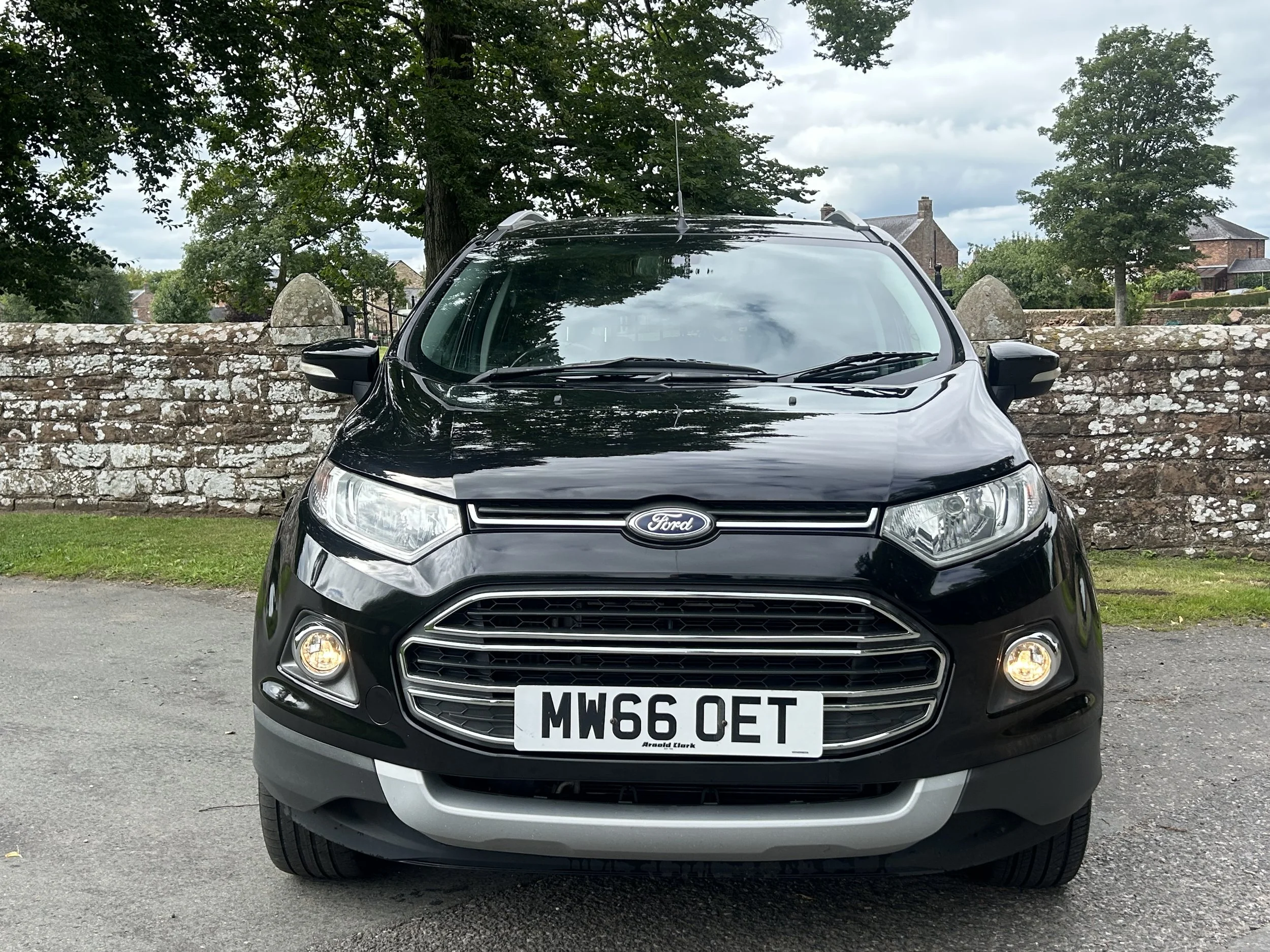 Front view of a black Ford SUV with UK license plate parked on a paved area, stone wall, grass, trees, and houses in the background.
