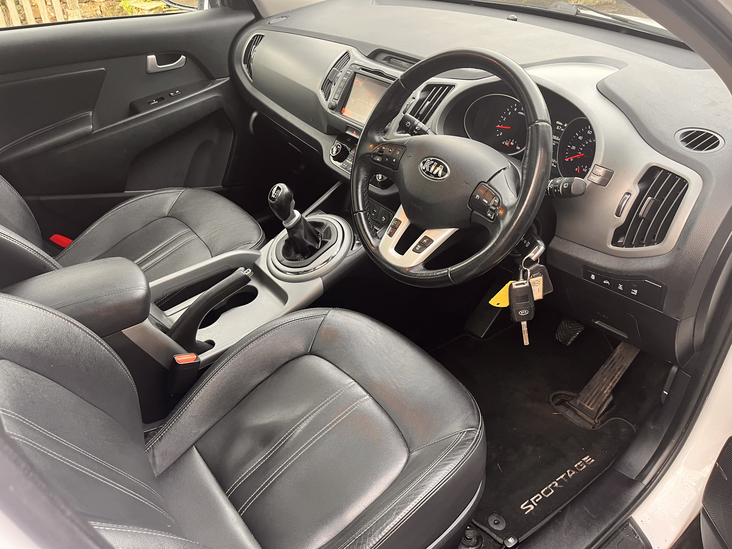 Interior of a Kia Sportage with black leather seats, manual gear shift, steering wheel with controls, dashboard with gauges, and key in ignition.