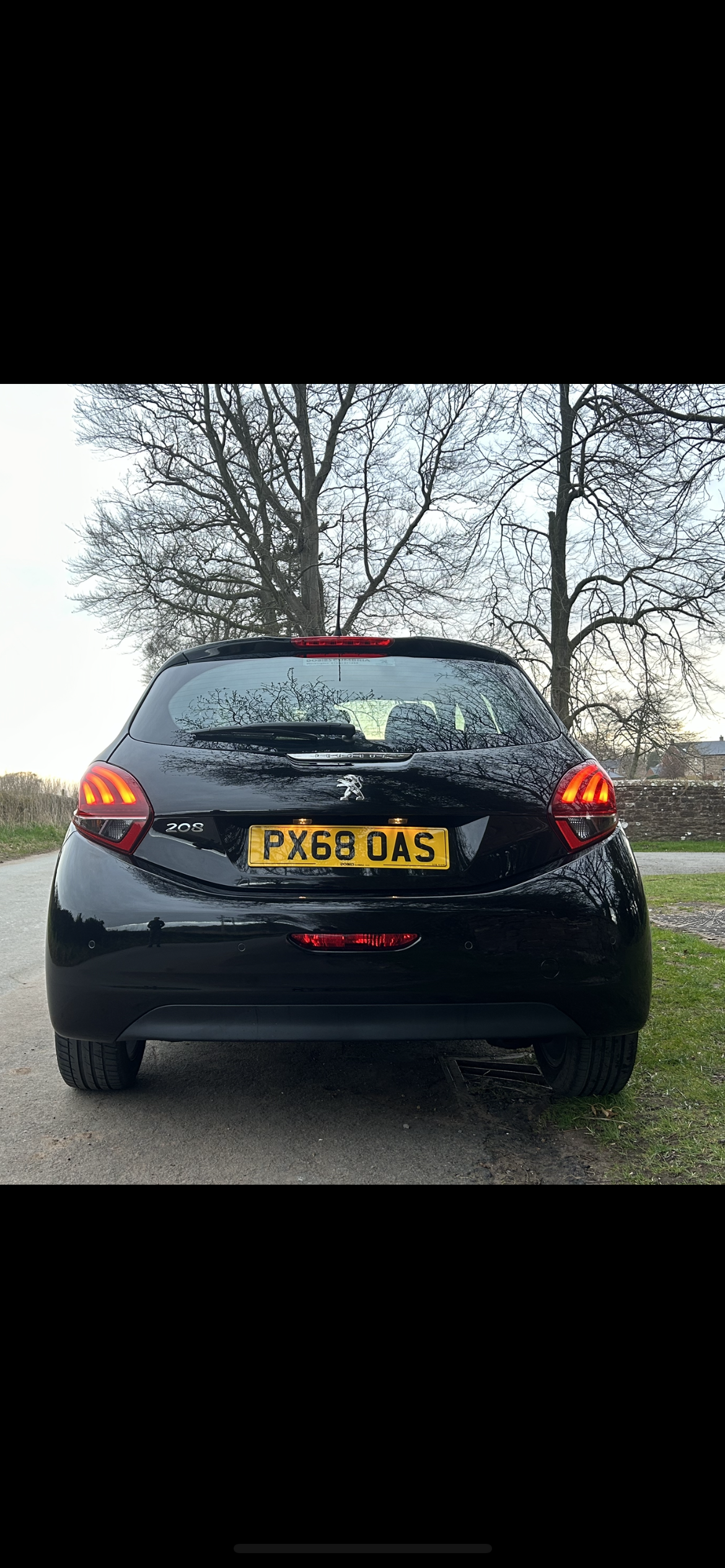 Rear view of a black Peugeot 208 with license plate PX68 OAS parked on a rural road, surrounded by bare trees.