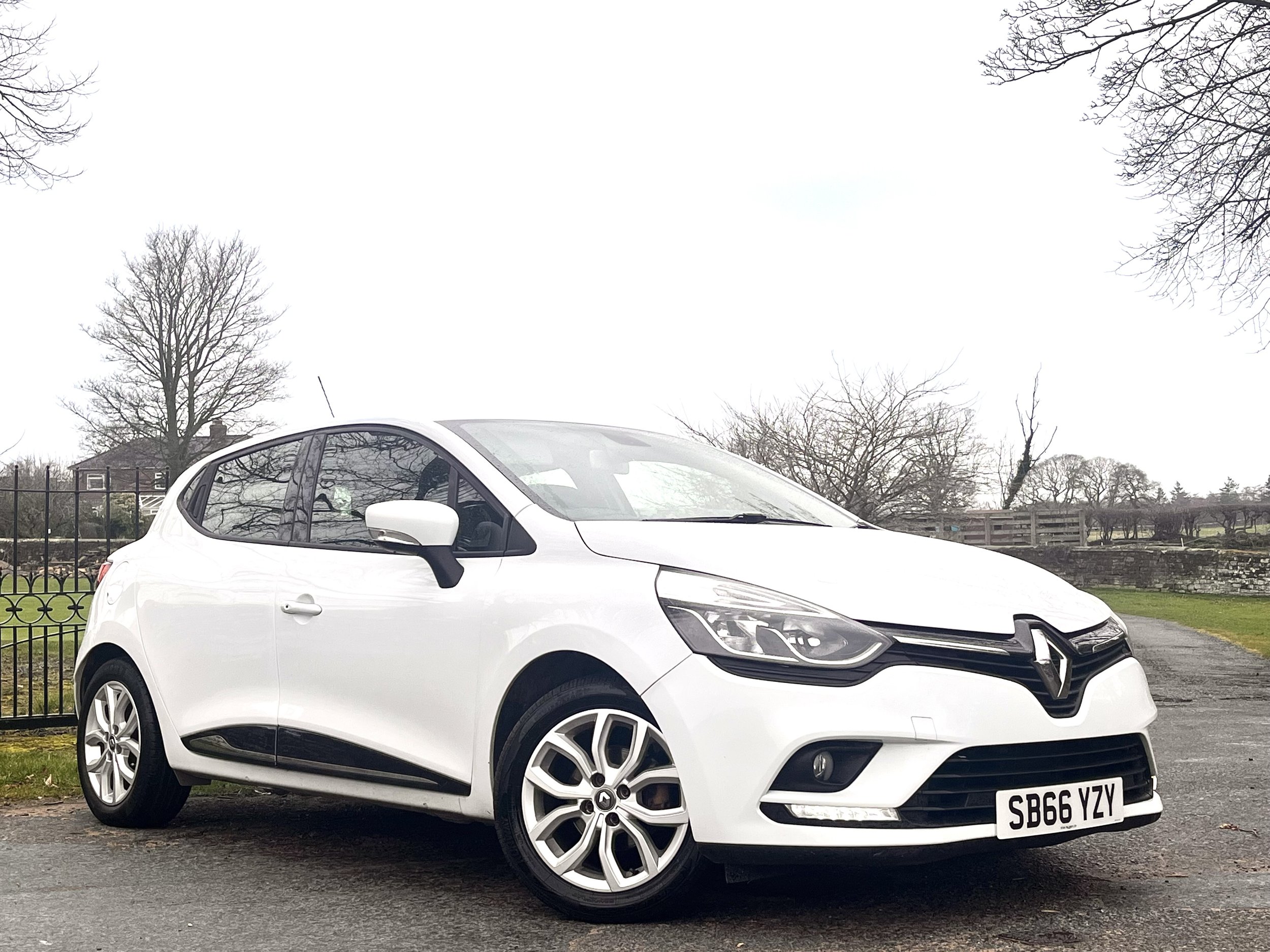 White Renault hatchback car parked on the side of a paved road with trees and a fence in the background.
