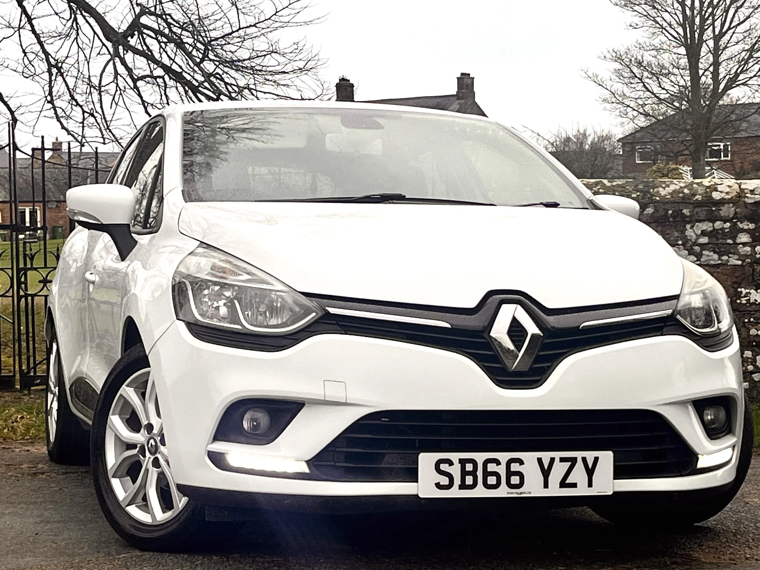 White Renault car parked on a street with a stone wall and residential houses in the background.