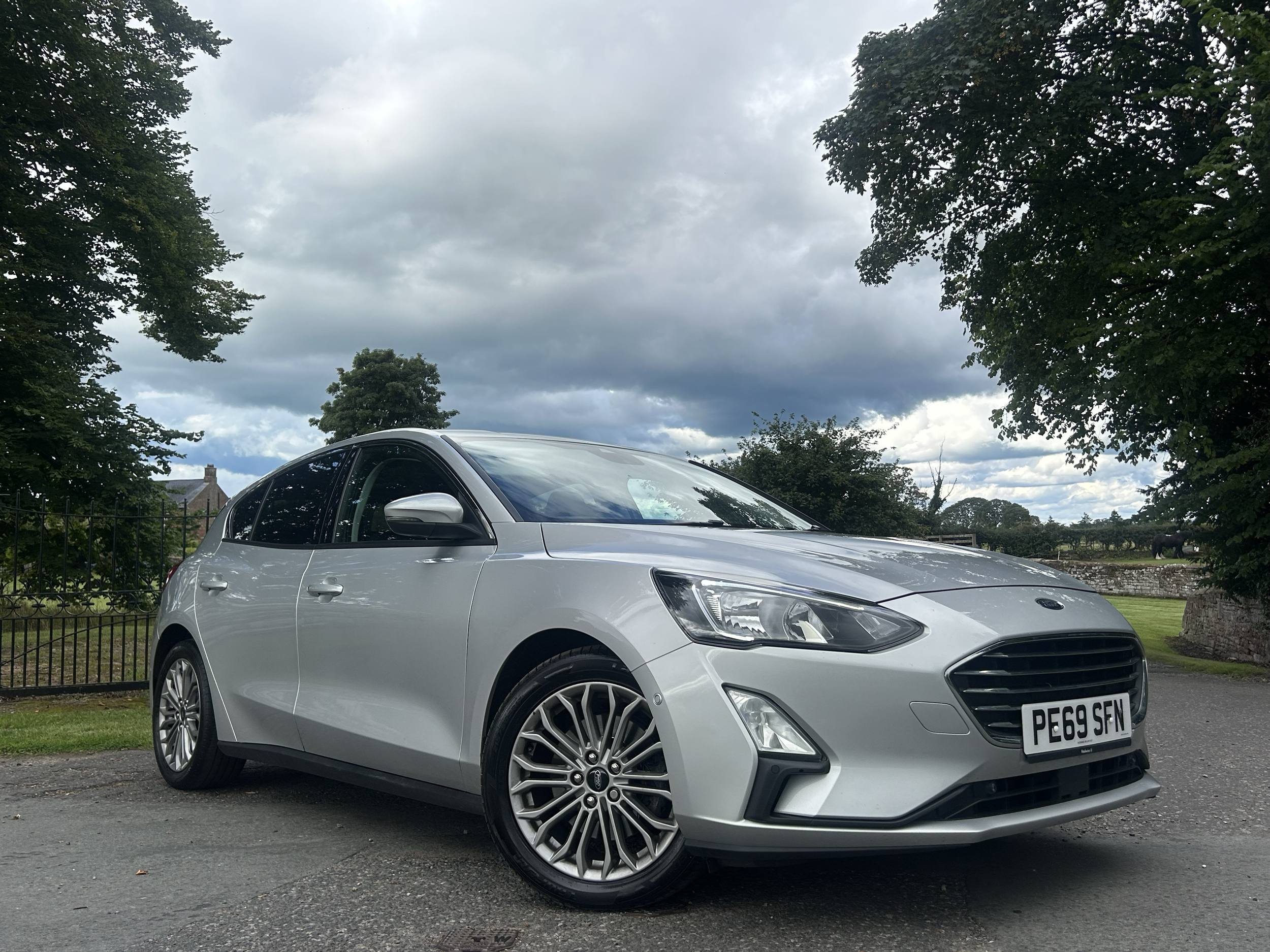 A silver Ford Focus parked on a paved surface with a black fence, trees, and cloudy sky in the background.