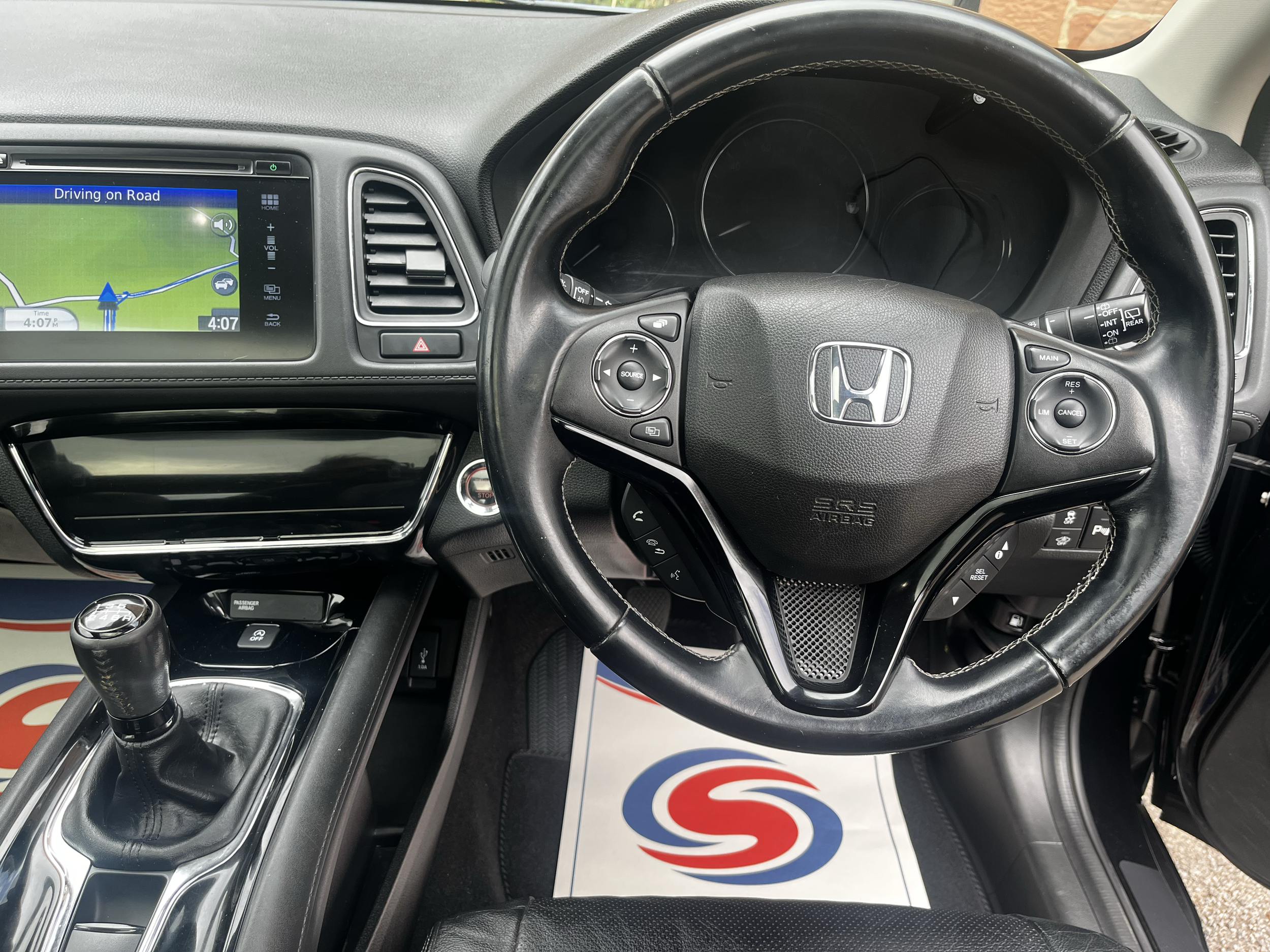 The image shows the interior of a car, specifically the driver's area, with a steering wheel featuring the Honda logo, a gear shift, and an infotainment screen displaying a GPS map and current time of 4:07 pm. The dashboard and air vents are also vis