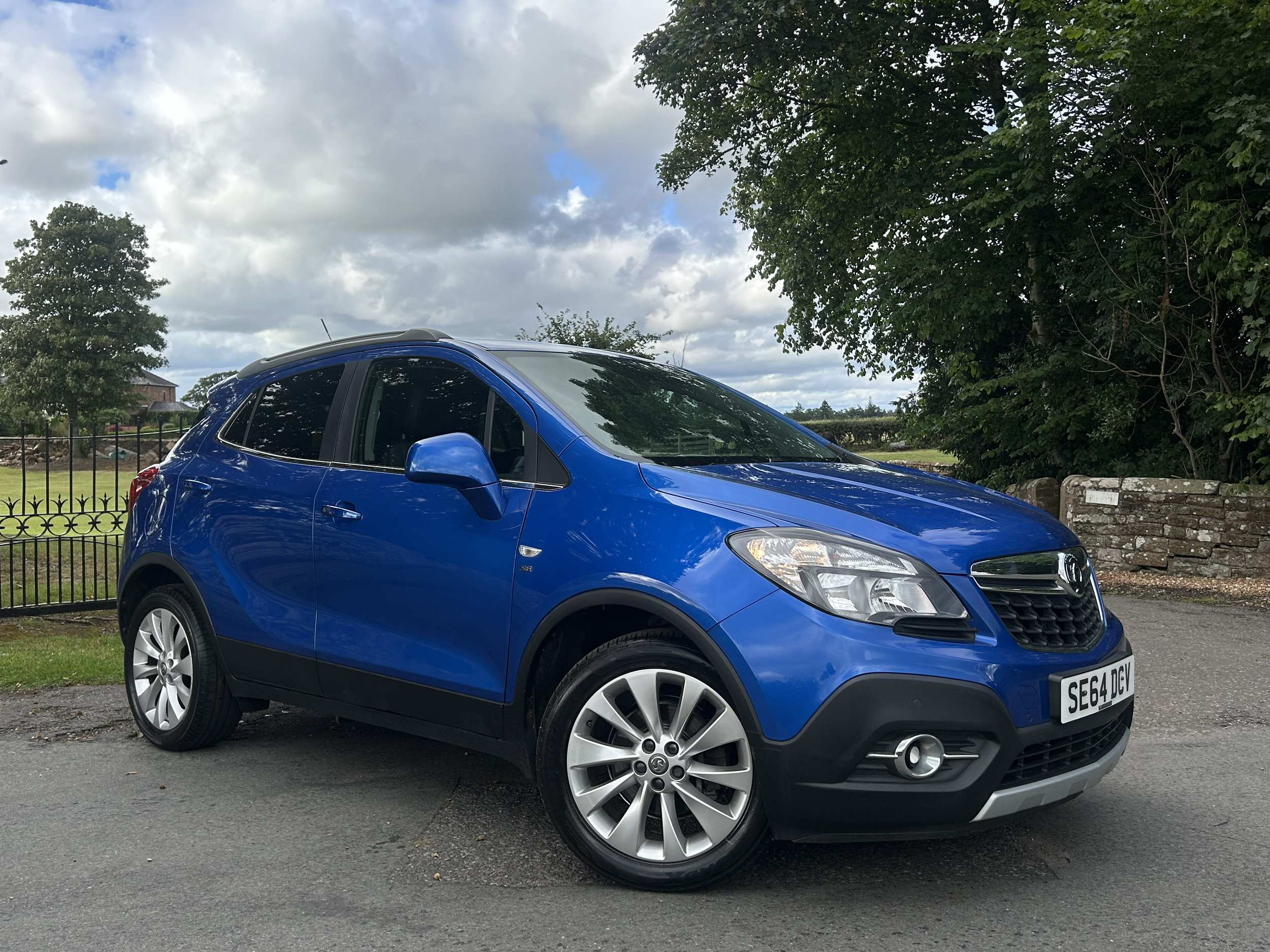 A bright blue compact SUV parked on a paved surface with trees and cloudy sky in the background.