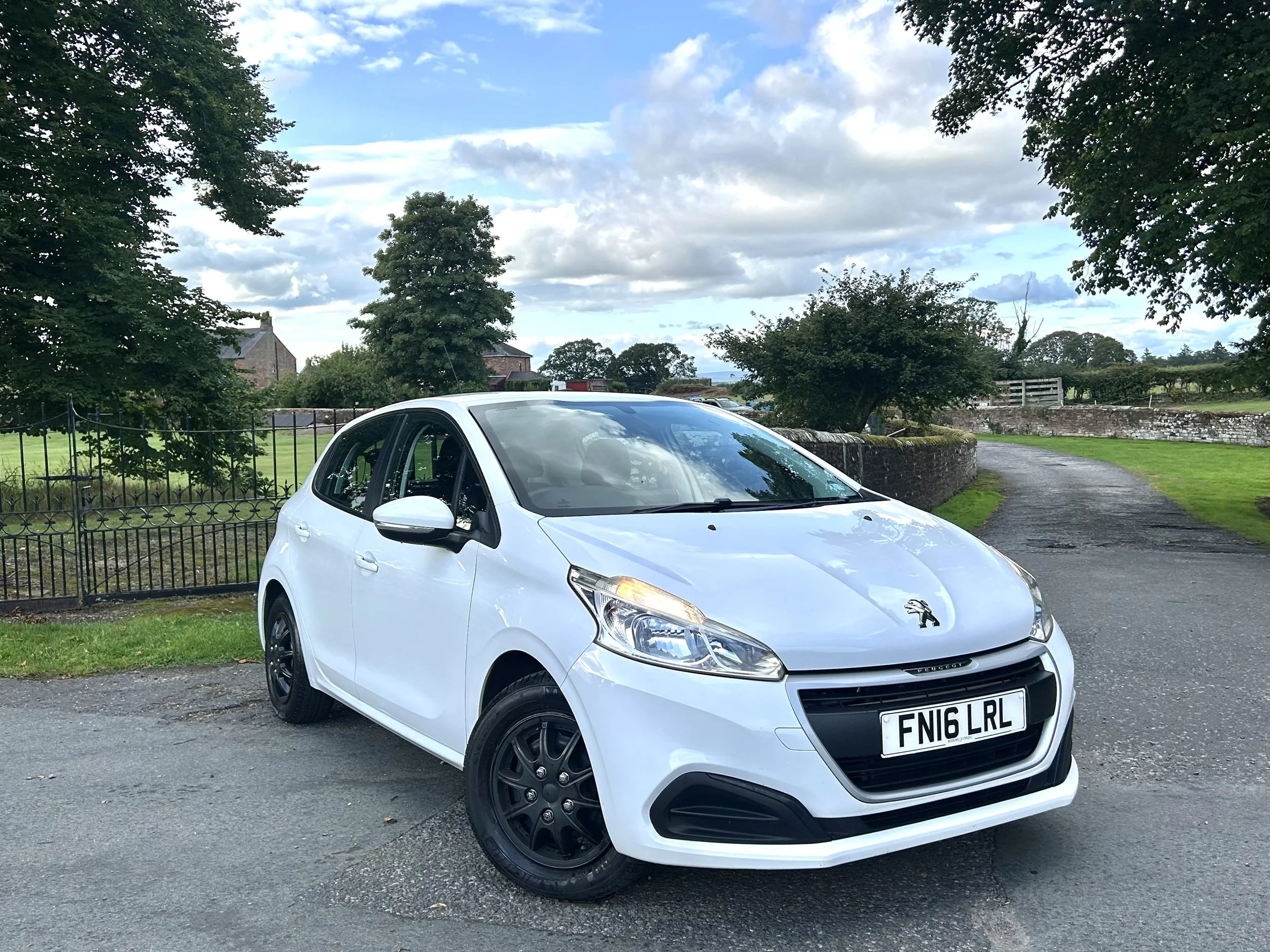 A white Peugeot car parked on a paved area near a grassy field, with a stone wall, trees, and houses in the background under a partly cloudy sky.