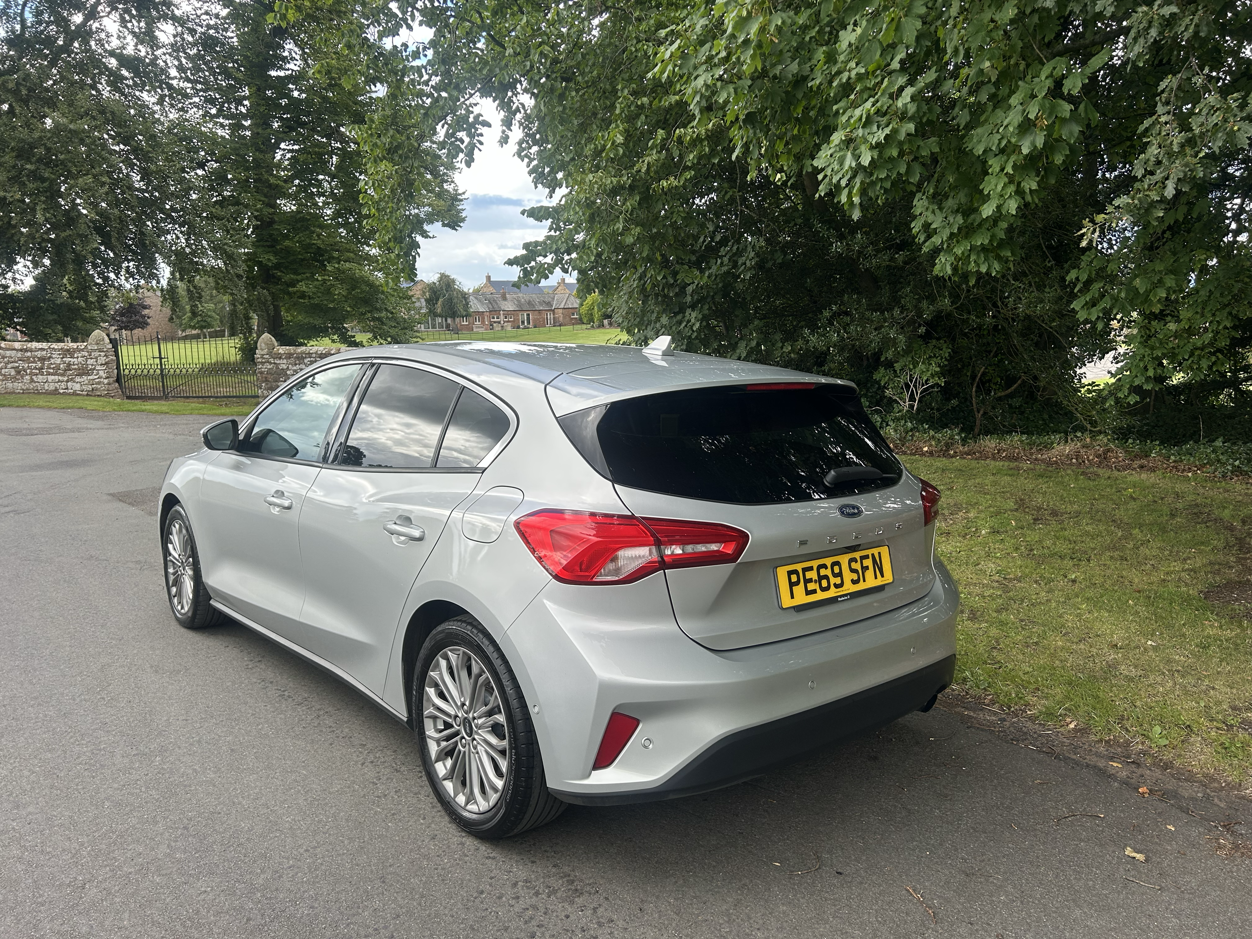 A silver Ford Focus hatchback car parked on a street next to a grass verge with trees and a stone wall with an ornate gate in the background.
