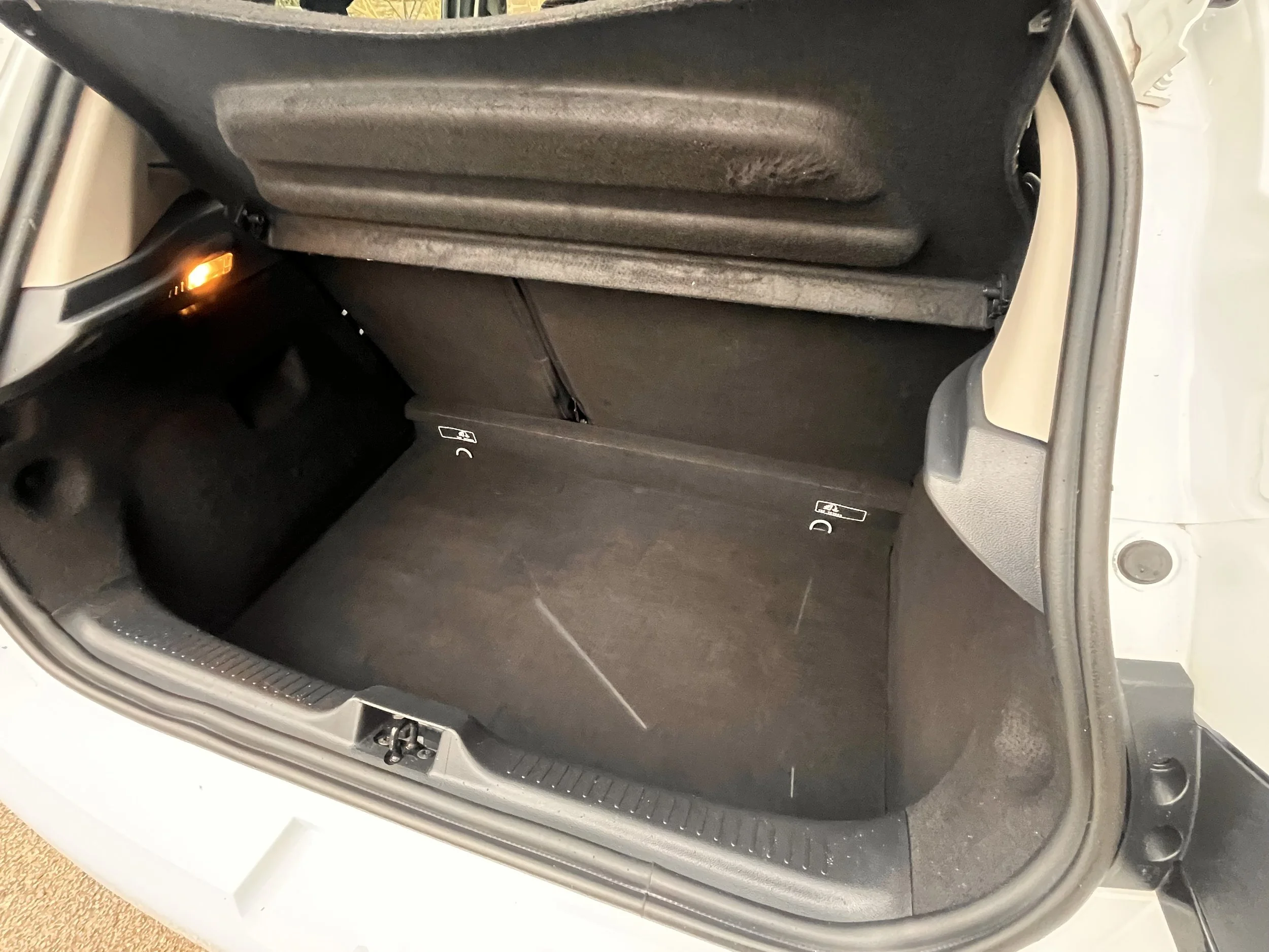 Interior of a car trunk with a black carpeted floor, some scratches, side compartments, and a small light on the left side outlined in orange.