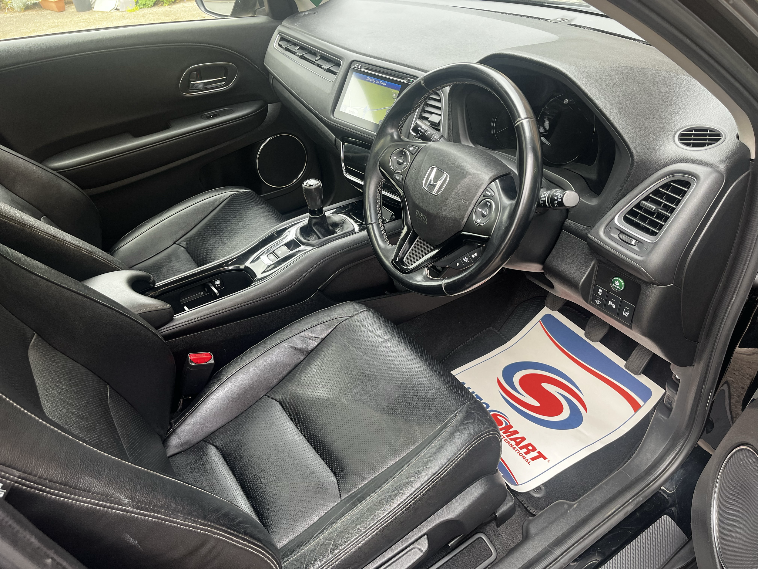 Interior of a Honda vehicle showing the steering wheel, dashboard, gear shift, and front seats with black leather upholstery. There is a screen on the dashboard and a floor mat with the logo of a car dealership.