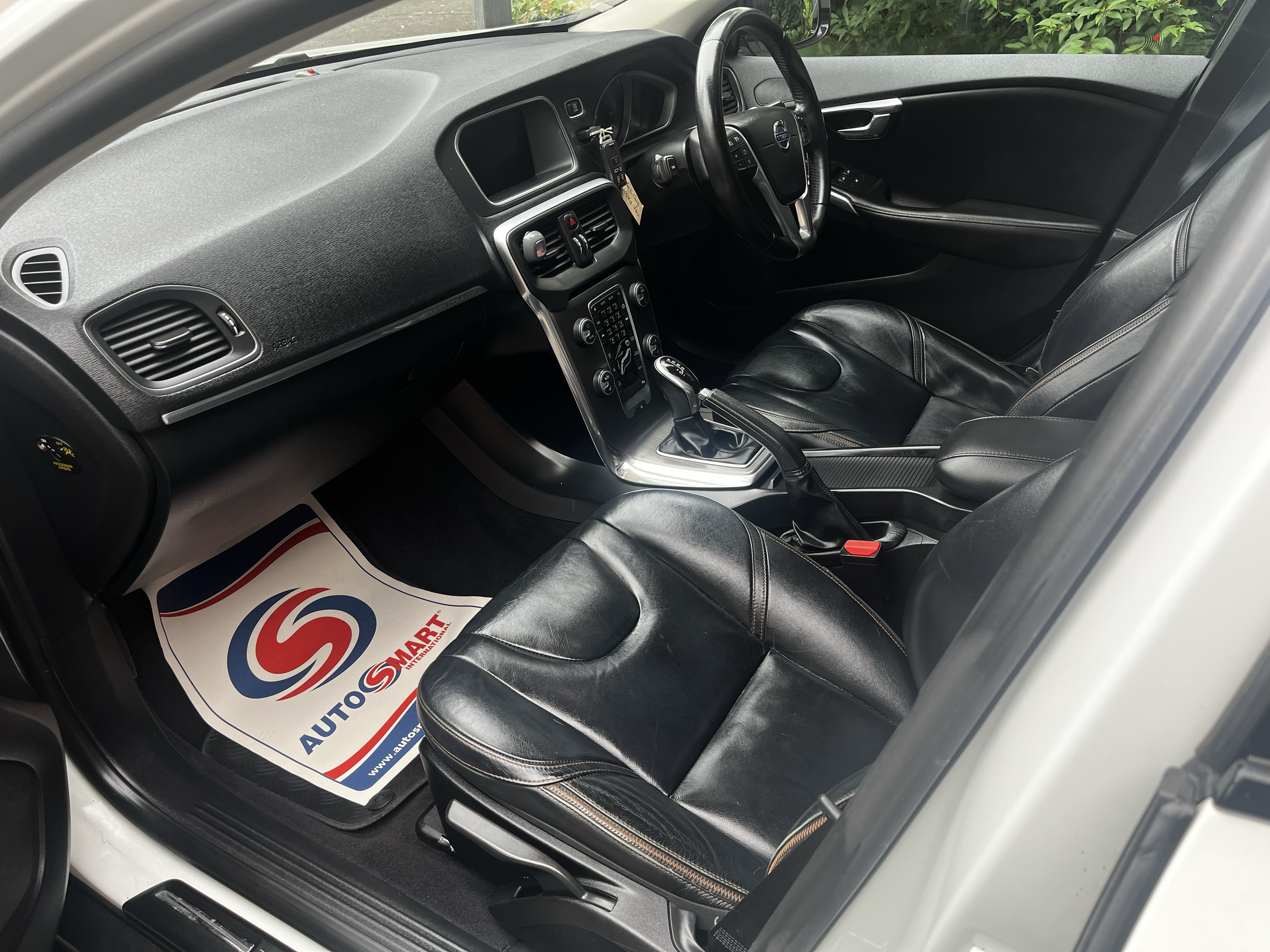 Interior of a car showing the dashboard, steering wheel, and front seats. There is a dealership floor mat on the driver side floor with the logo and website of Auto Smart.