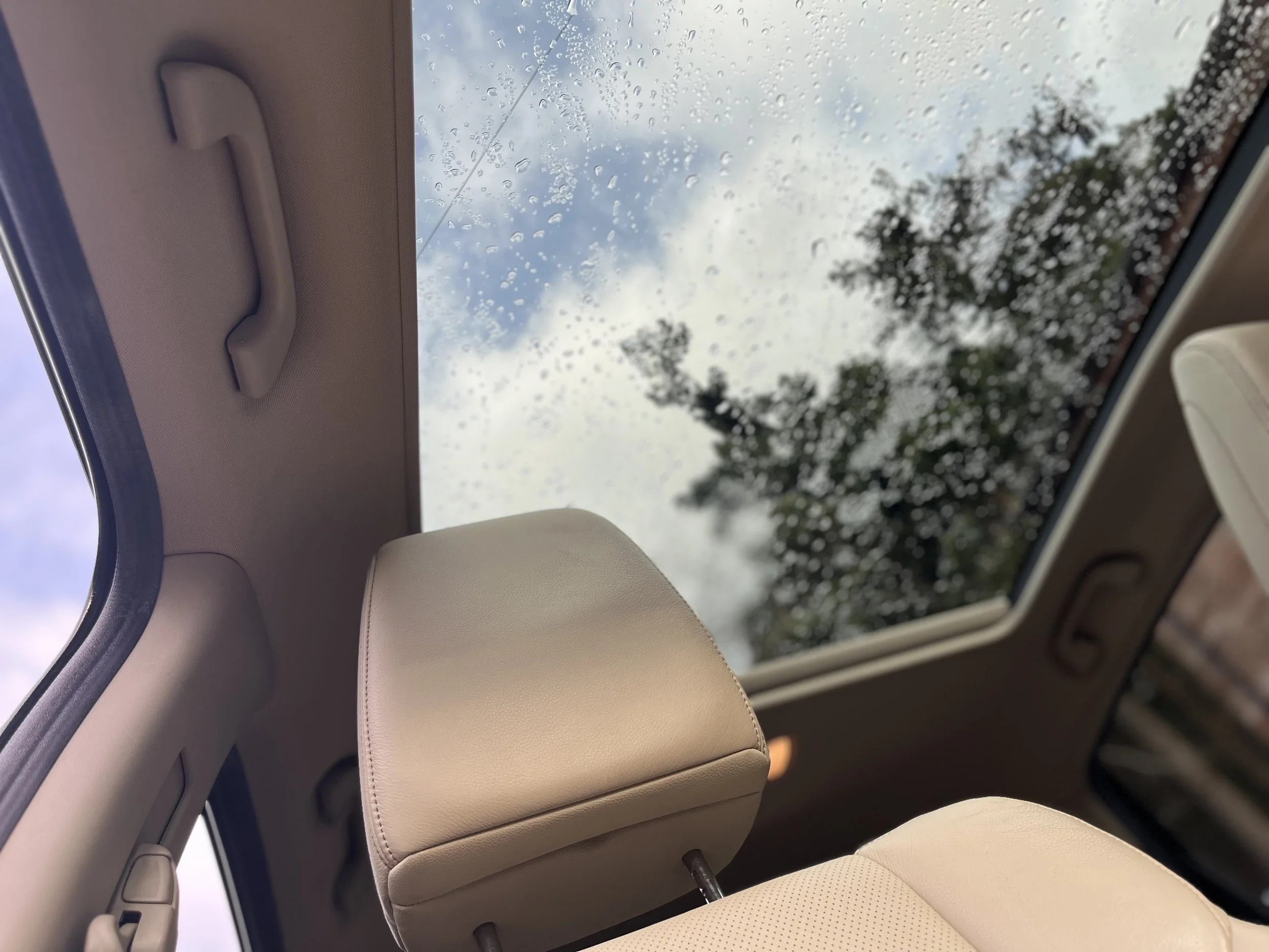 Interior view of a car's back seat showing a beige headrest and a large rear window with raindrops on it, through which trees and a cloudy sky are visible.