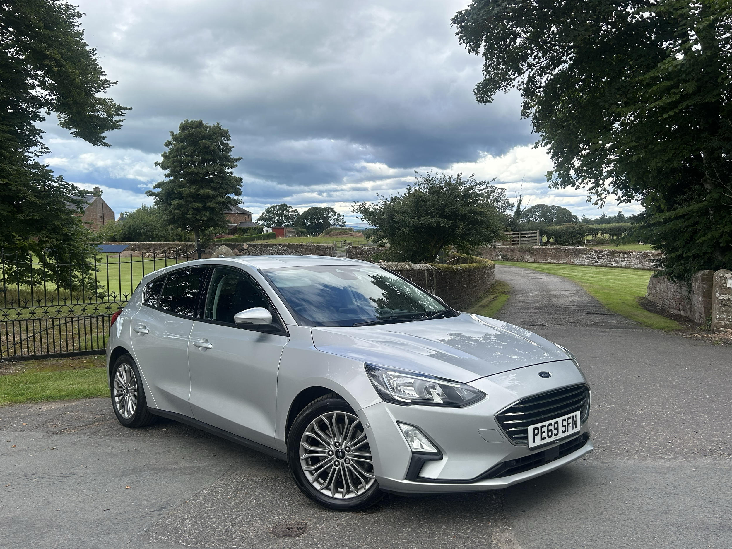 A silver hatchback car parked on a paved area near a rural scene with trees, a stone wall, and small buildings under a cloudy sky.