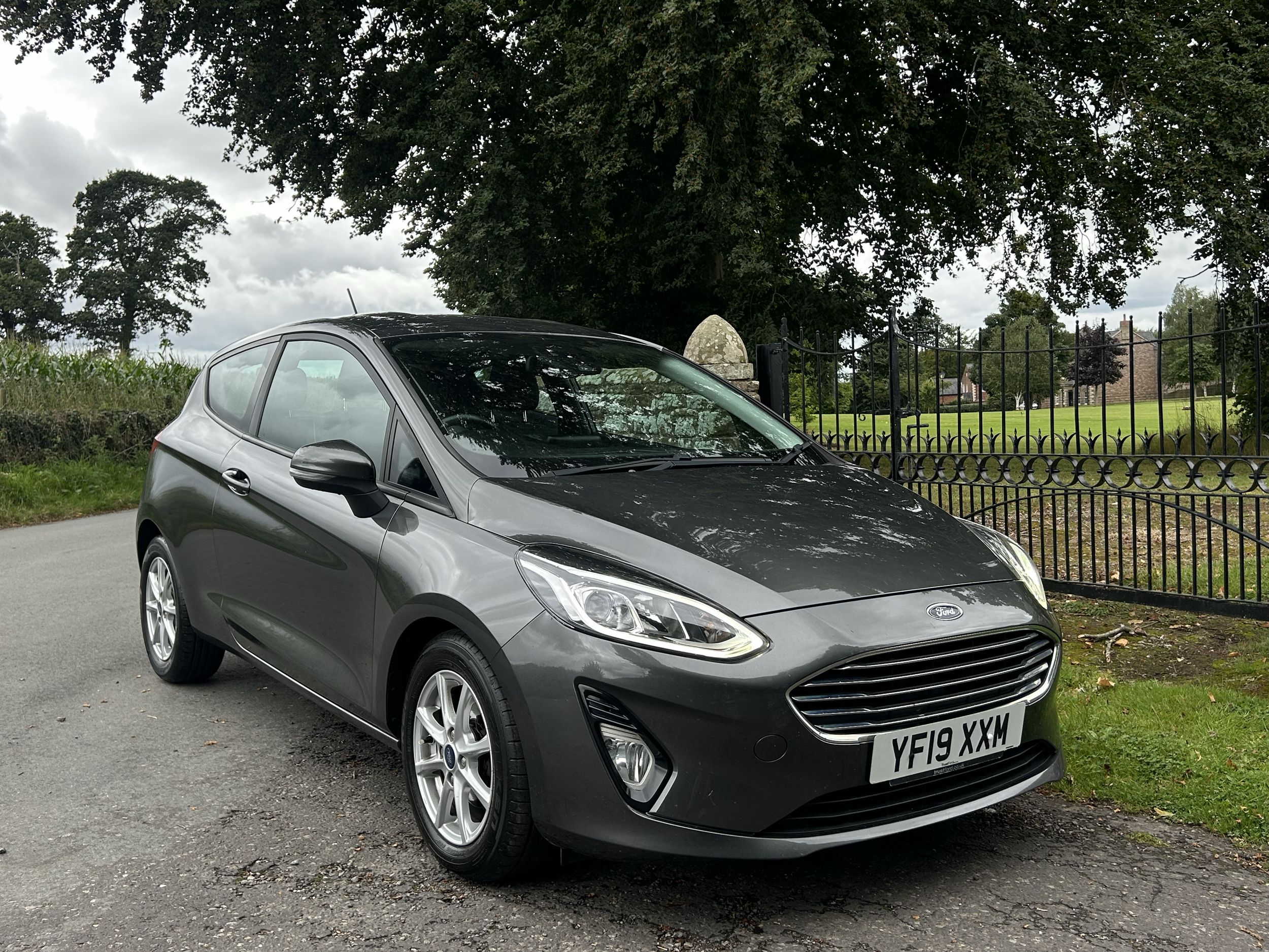 A black Ford car parked on a pavement with green grass and trees in the background, surrounded by a black metal fence.