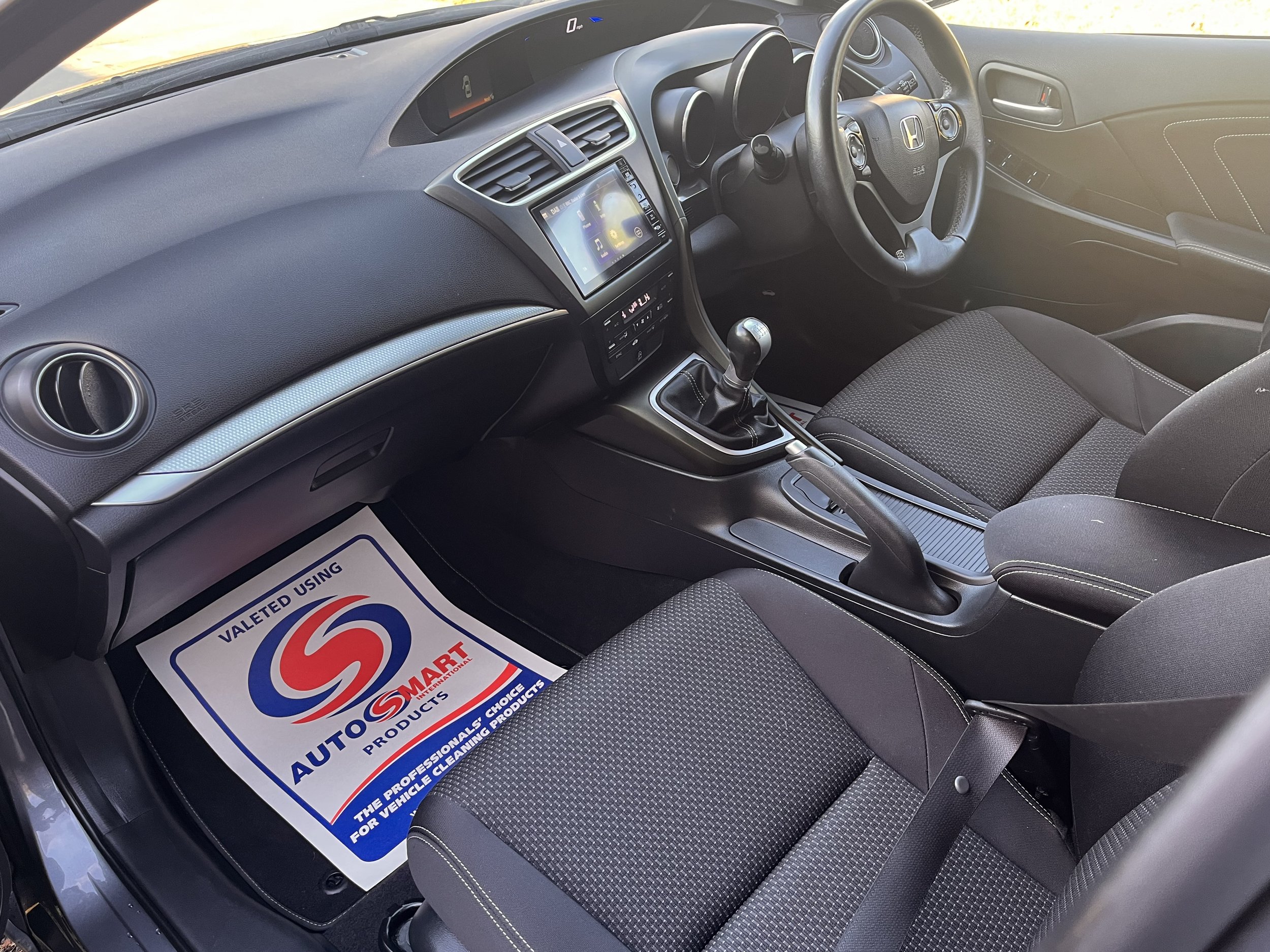Interior of a Honda car showing the dashboard, steering wheel, gear shift, and front seats. A floor mat with 