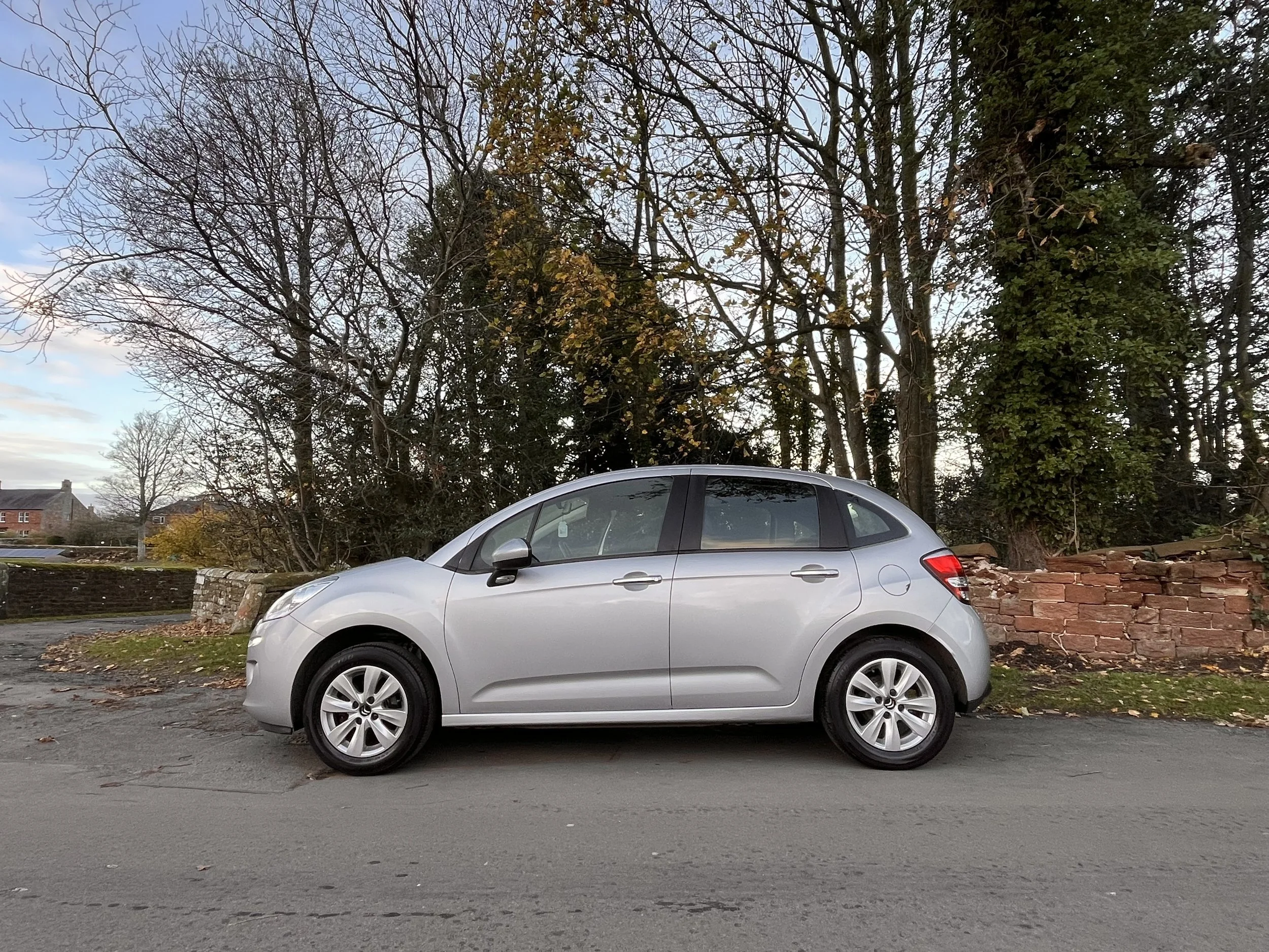 A silver hatchback car parked on a street with trees and a brick wall in the background, autumn leaves on the ground.