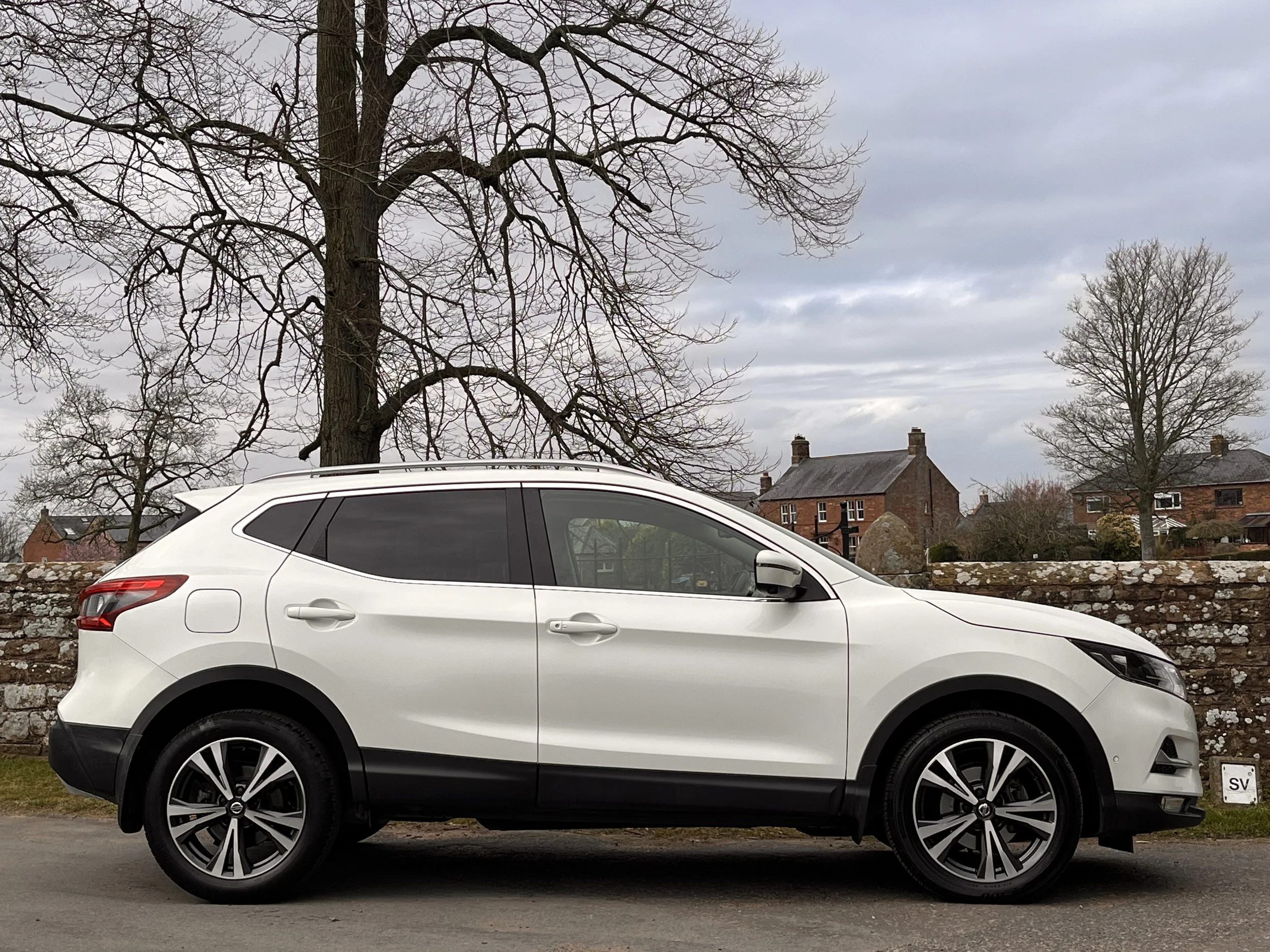 White SUV parked along a street with a stone wall, leafless trees, and brick houses in the background under a cloudy sky.