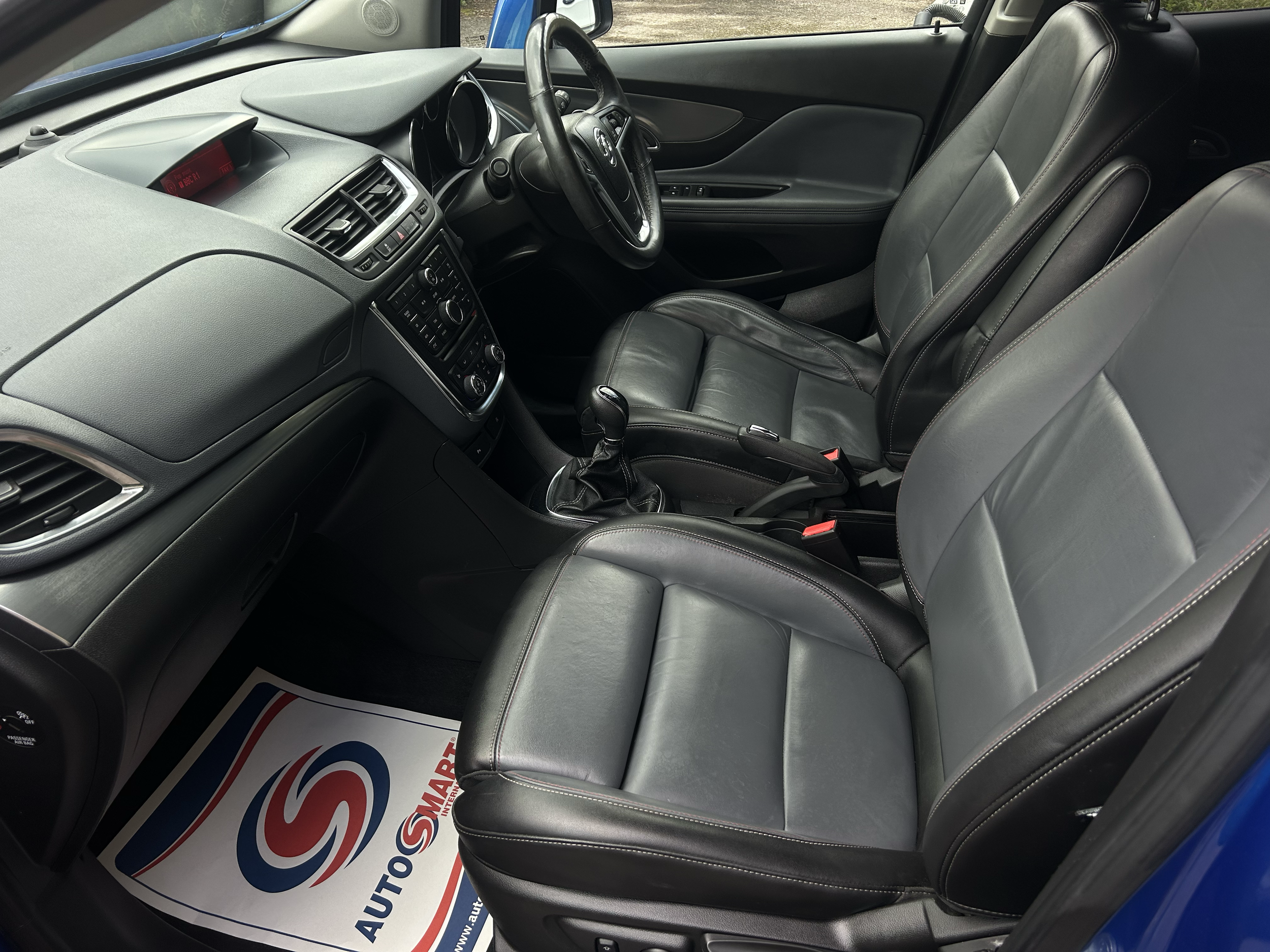 The interior of a car showing black leather front seats, dashboard, steering wheel, gear shift, and a paper mat with a logo on the floor.