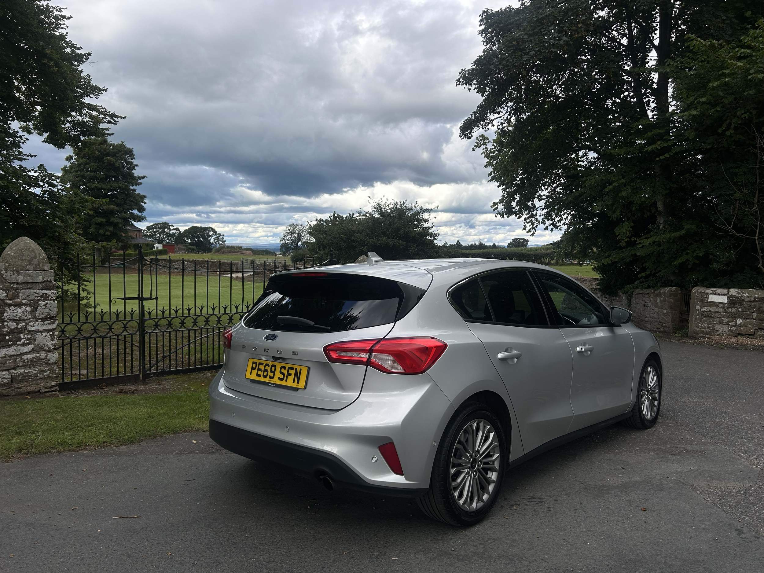 A silver Ford hatchback car parked on a driveway with a background of trees, a stone wall, a gated fence, and overcast clouds in the sky.